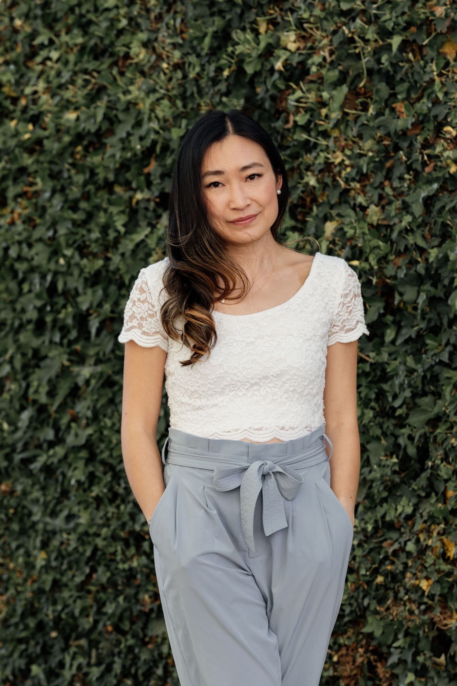 Young Asian woman in white lace crop top and gray bow-tie trousers posing against a lush green ivy wall.