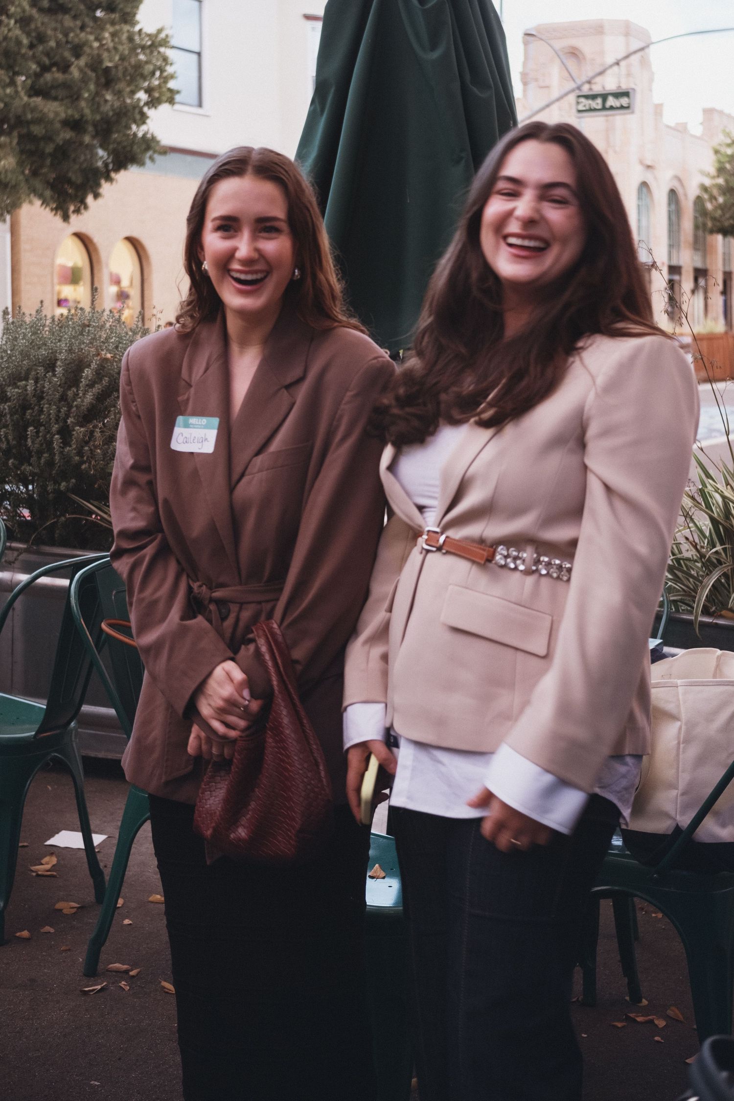 Two young women in blazers laughing together outdoors at a casual event, wearing name tags.