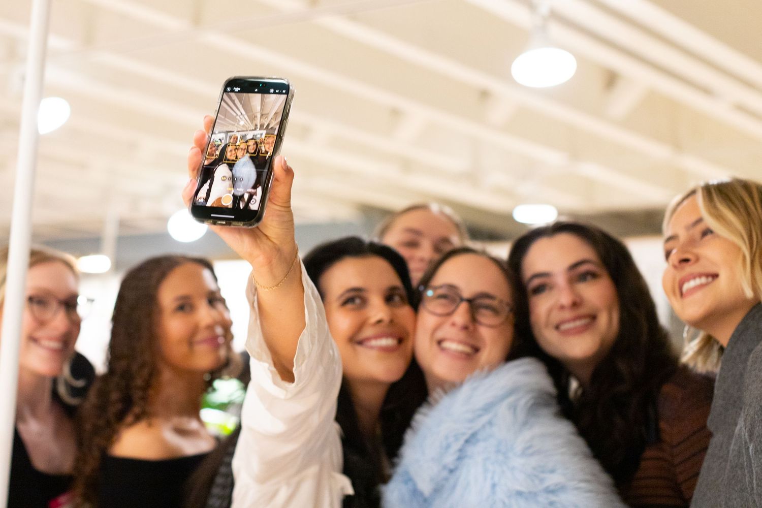 Group of smiling women taking a fun selfie together with a smartphone at an indoor event.
