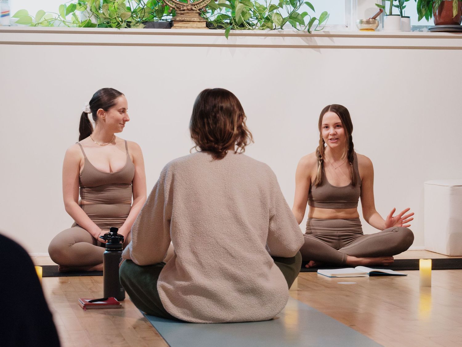 Three women in brown activewear sit cross-legged on yoga mats in a bright studio, engaged in a mindful group discussion.