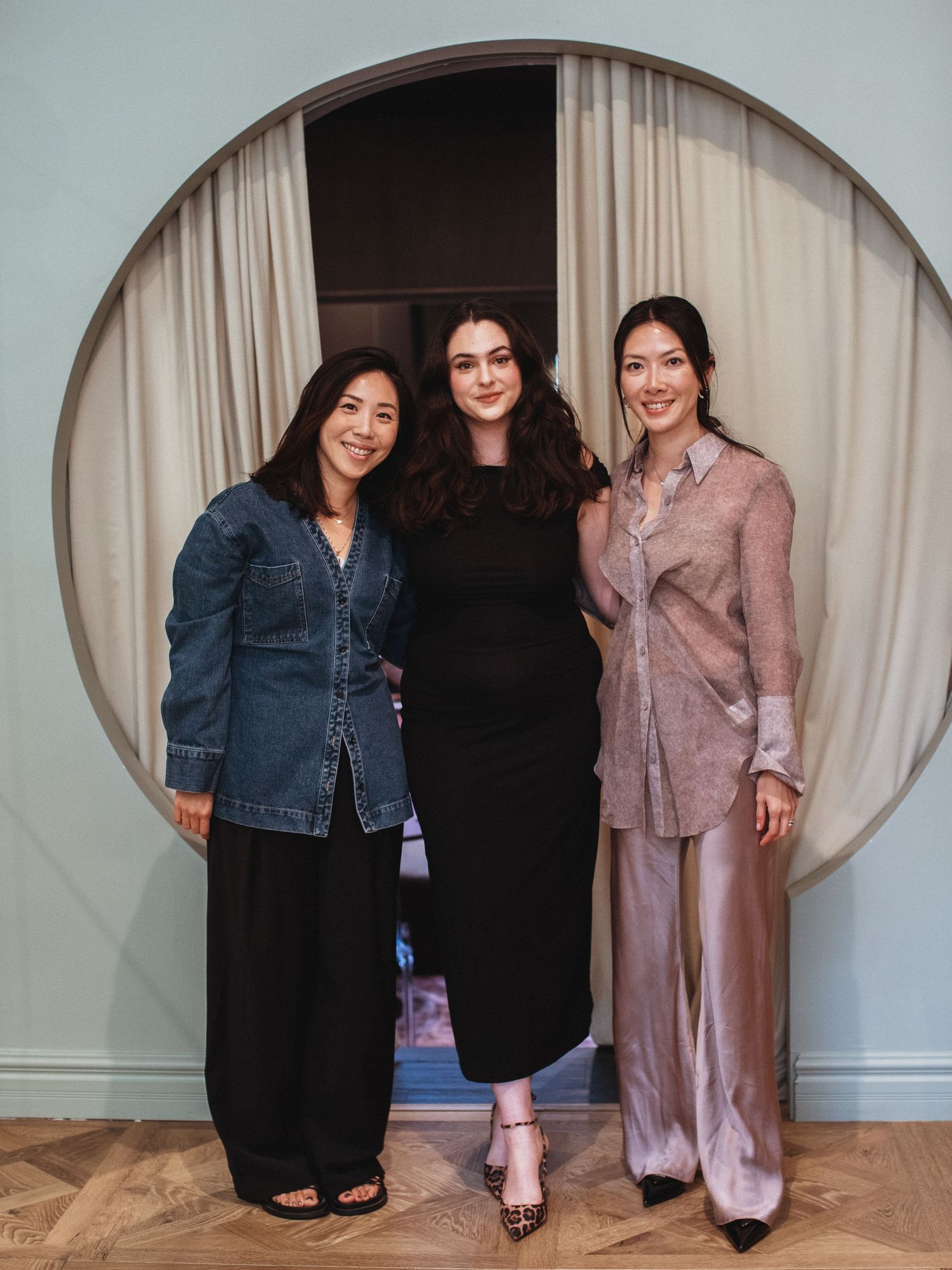 Three women smiling together in front of a circular arch doorway with neutral curtains in an elegant interior.