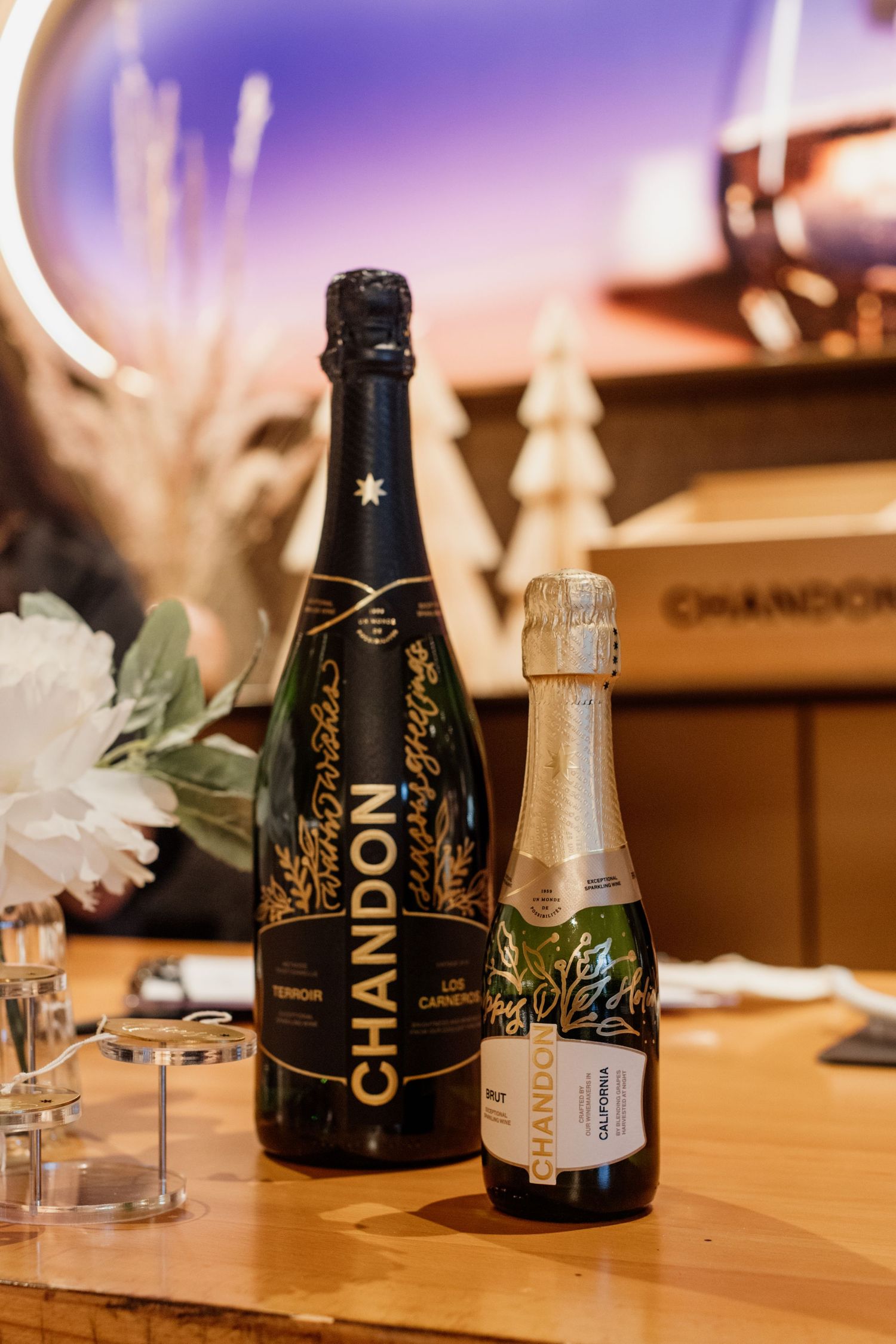 Two Chandon sparkling wine bottles displayed on a wooden table with festive holiday decorations in the background.