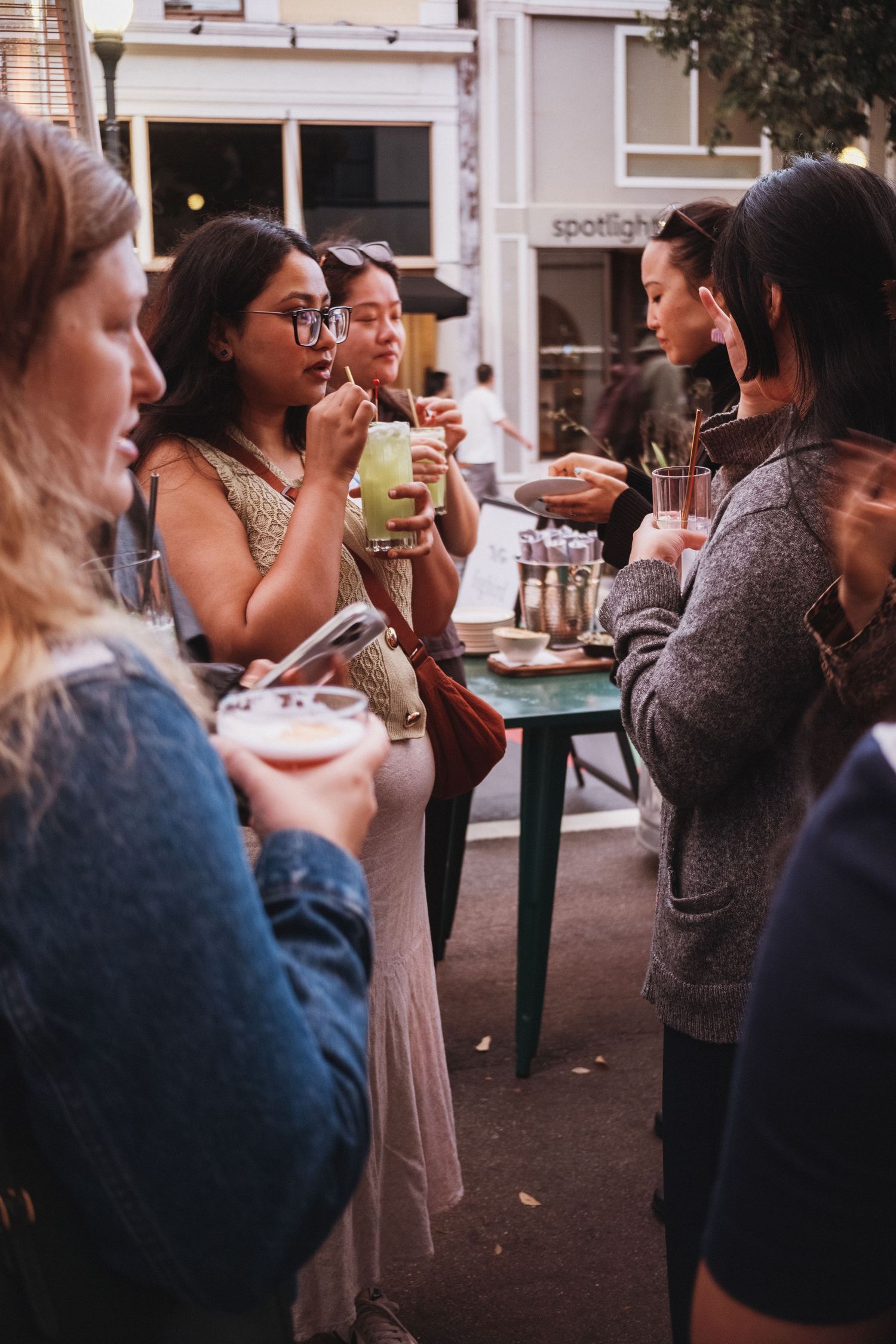 Group of women socializing outdoors at a street gathering, holding drinks and snacks near a cafe.