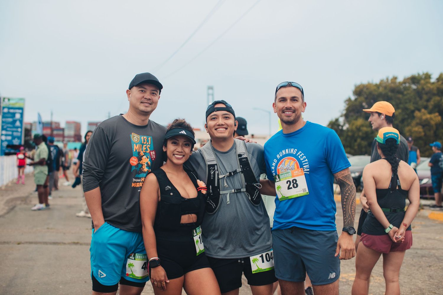 Four friends pose together smiling at an outdoor marathon race event on a cloudy day.