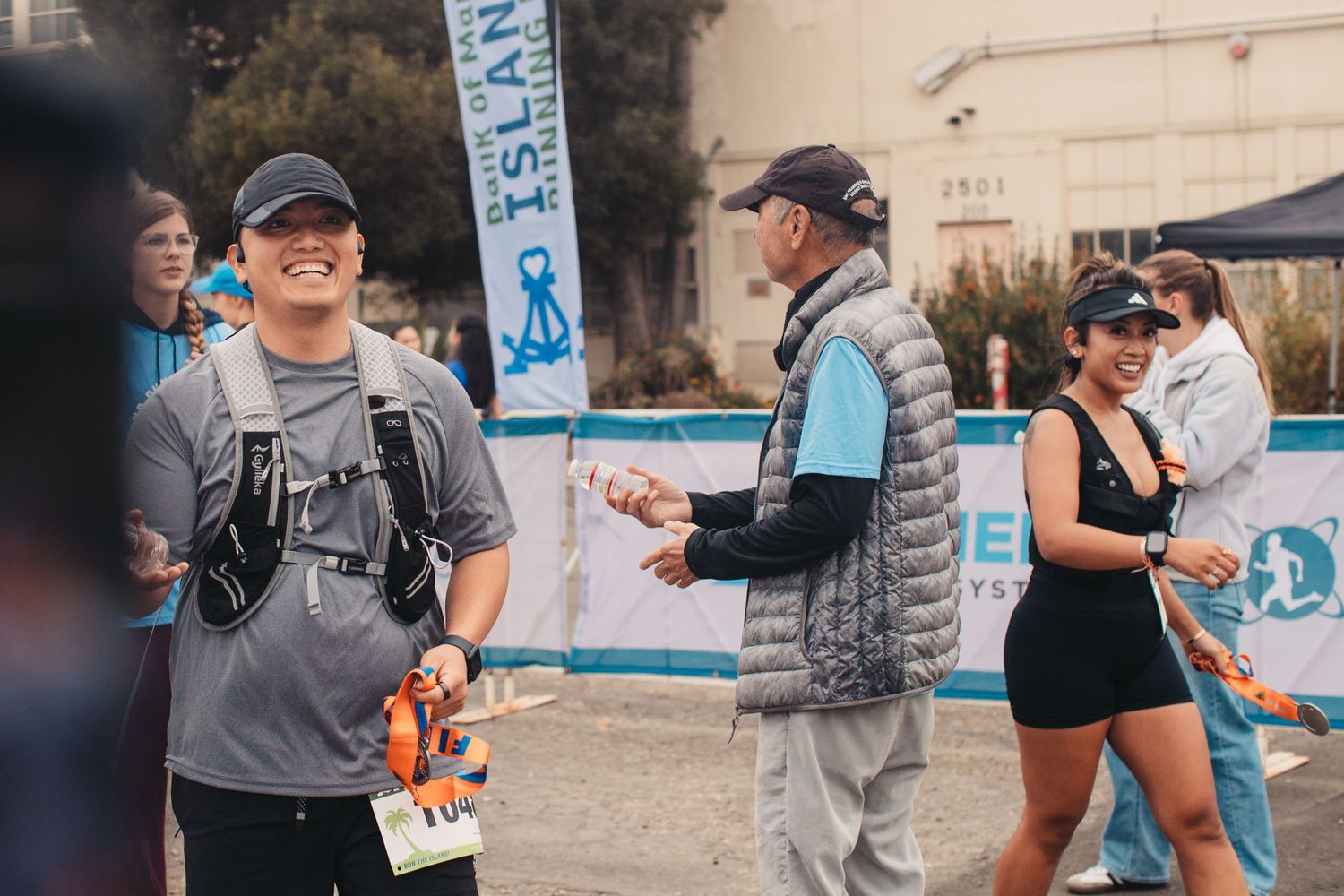 Runners smile at a finish line event near Alcatraz Island, wearing race vests and casual athletic wear outdoors.
