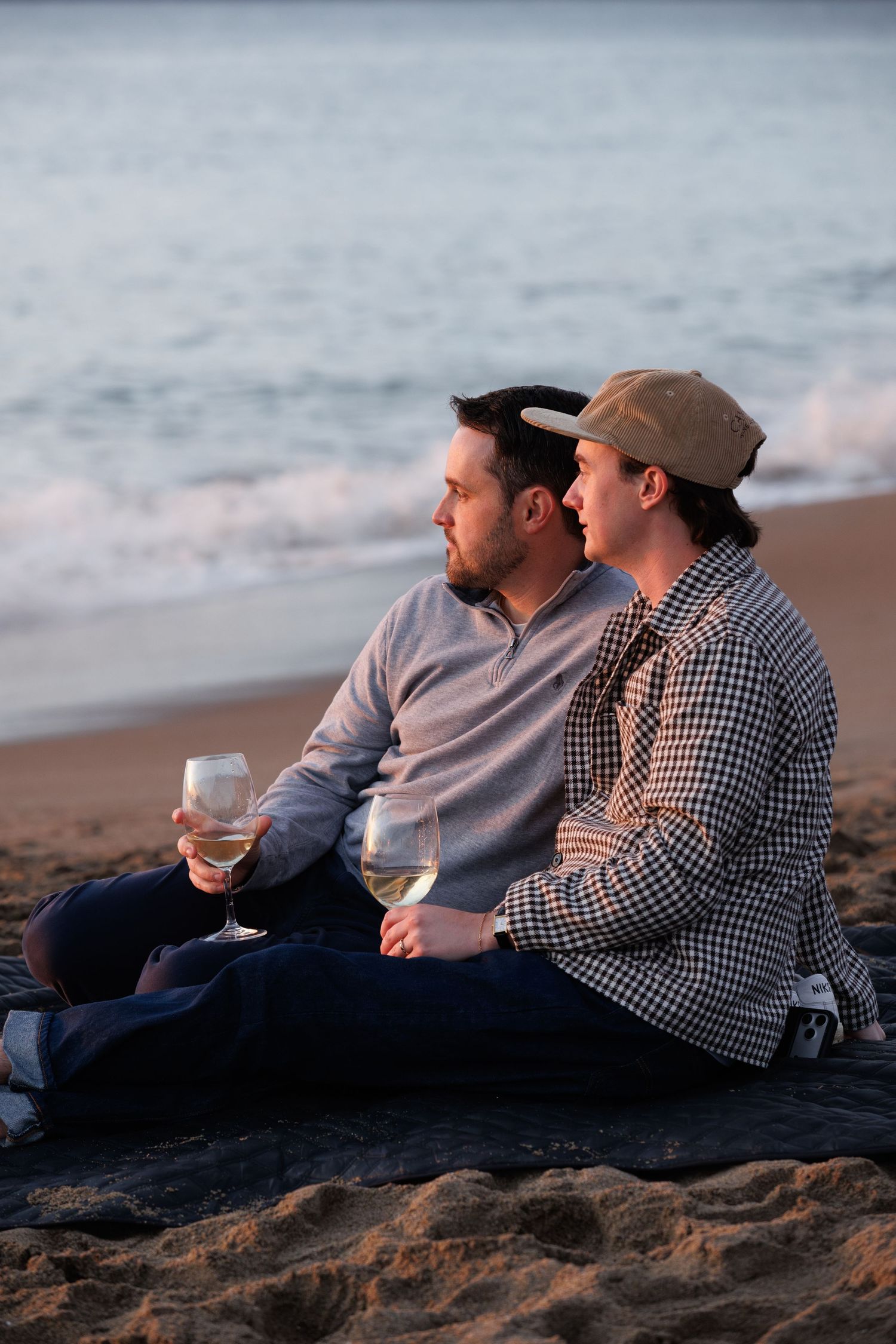 Two men sitting on a beach at sunset, sharing wine glasses and enjoying a romantic moment by the ocean waves.