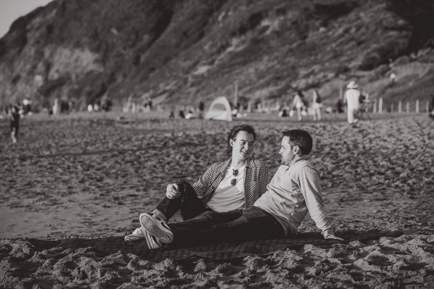 Black and white photo of a couple sitting together on a beach with rocky cliffs in the background.