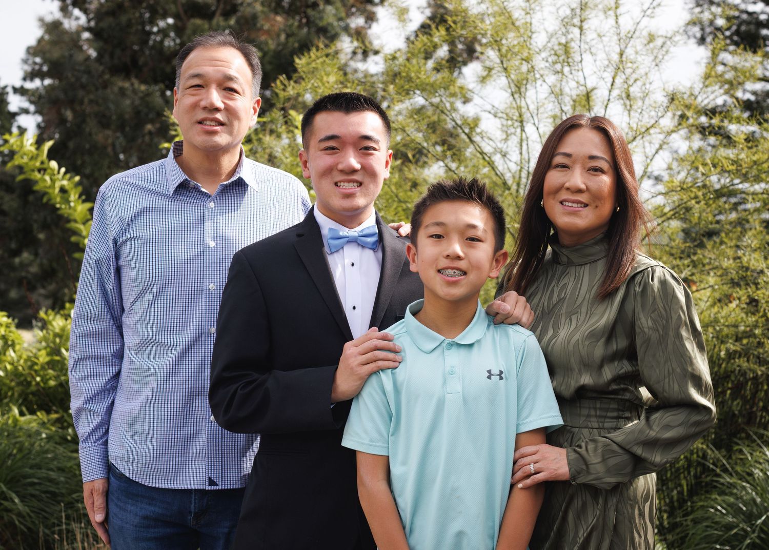 Family of four smiling outdoors, two adults and two boys, dressed in semi-formal attire with lush green trees behind.