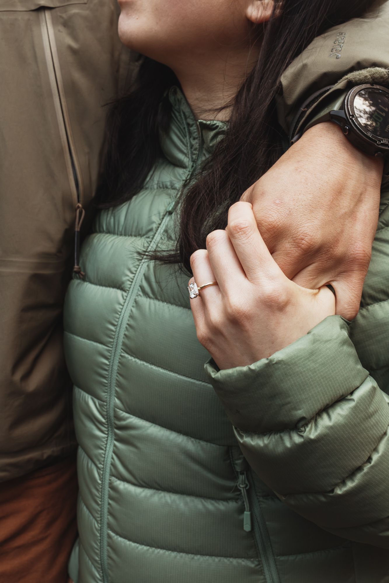 Couple embracing outdoors, woman in sage green puffer jacket holding mans arm, engagement ring visible on her finger.