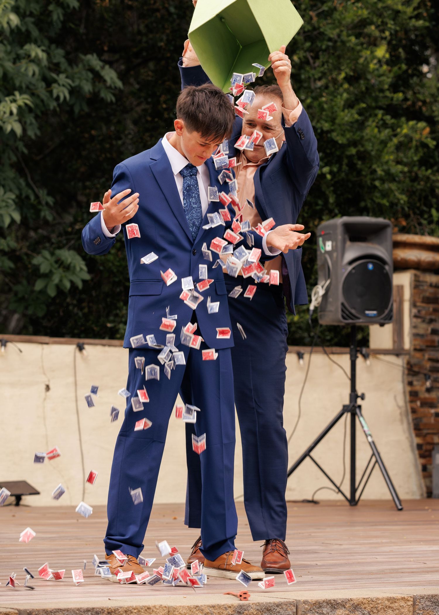 Two magicians in blue suits perform a dramatic playing card shower trick outdoors on a stage.