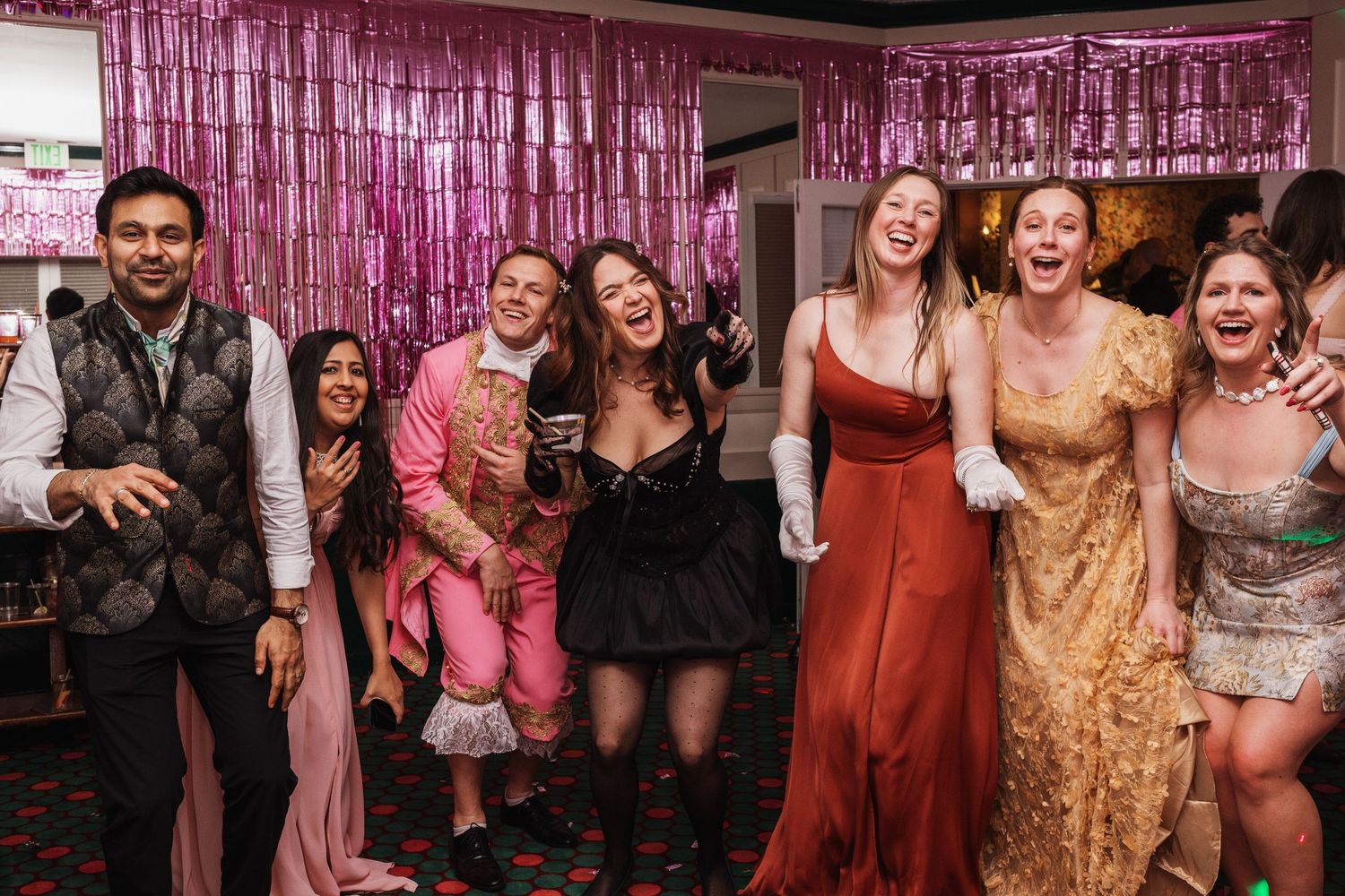 Group of friends in formal attire celebrating at a glamorous party with pink tinsel backdrop.