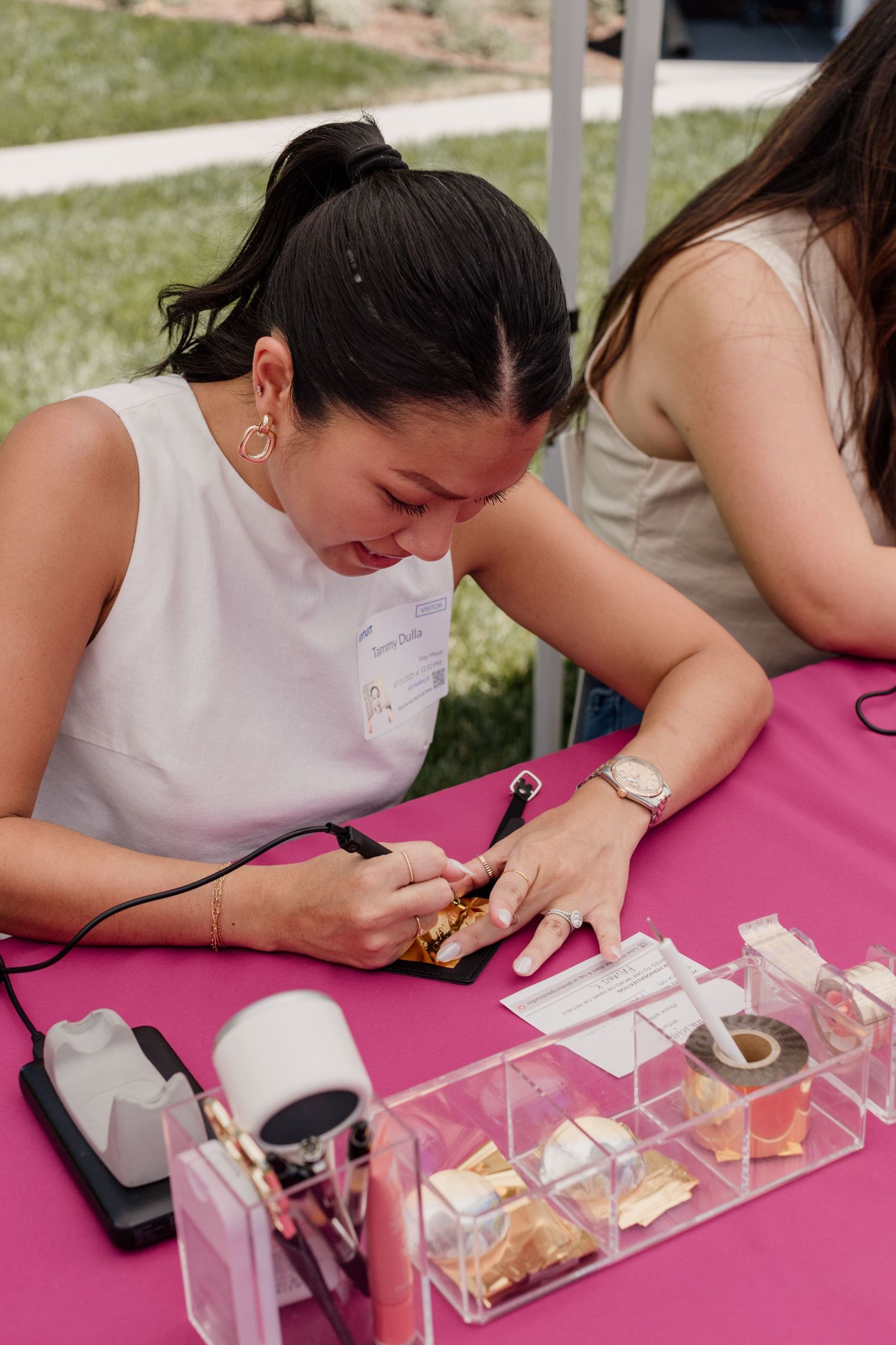 Woman doing nail art at a pink table outdoors at a beauty event with nail supplies displayed.