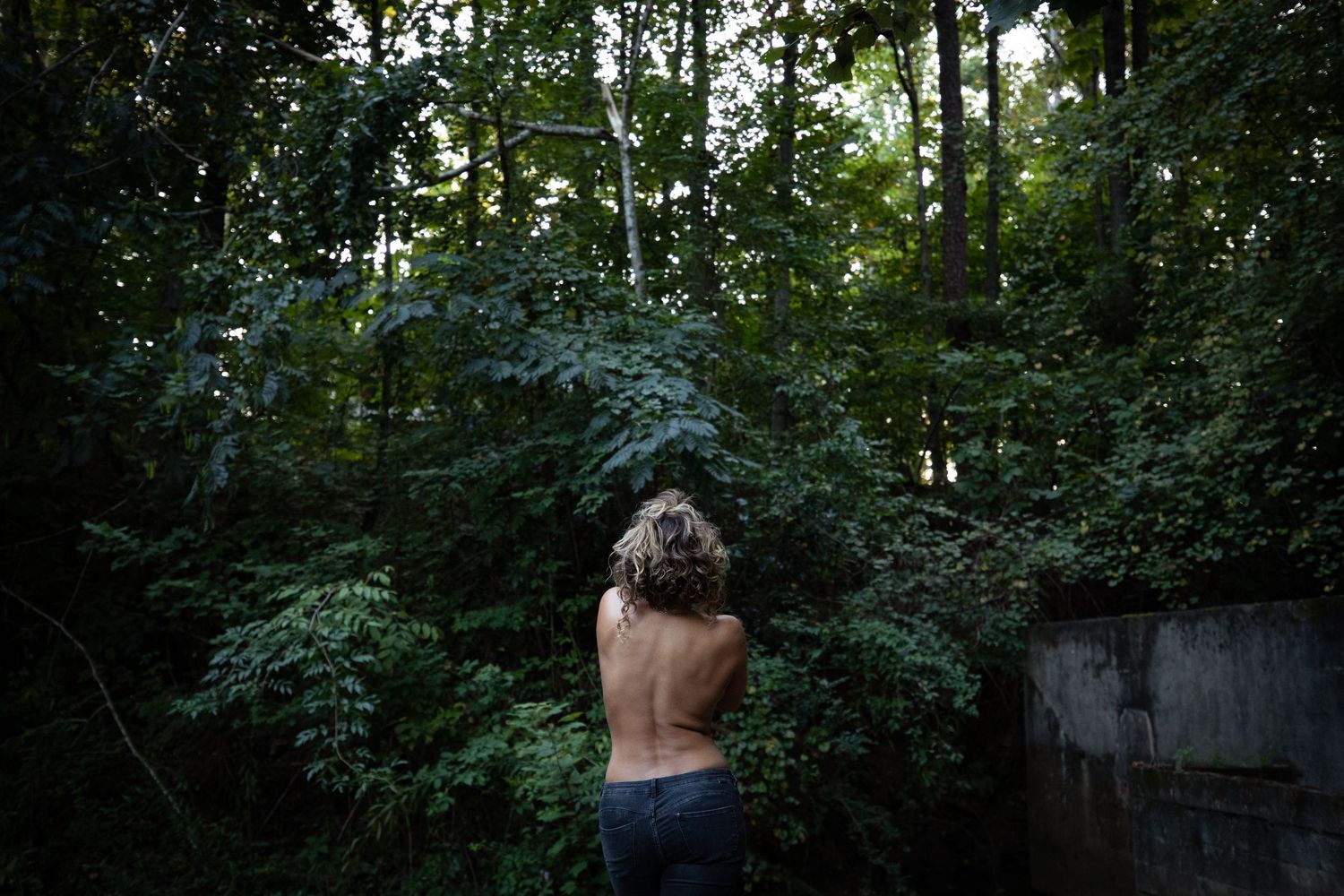 A person stands shirtless in front of lush green forest foliage on a summer evening.