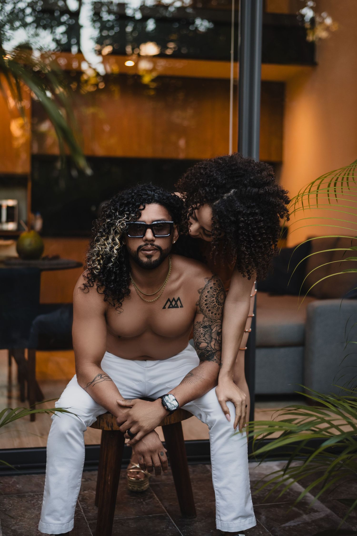 A stylish couple sharing an intimate moment on a patio with tropical plants and warm lighting in the background.