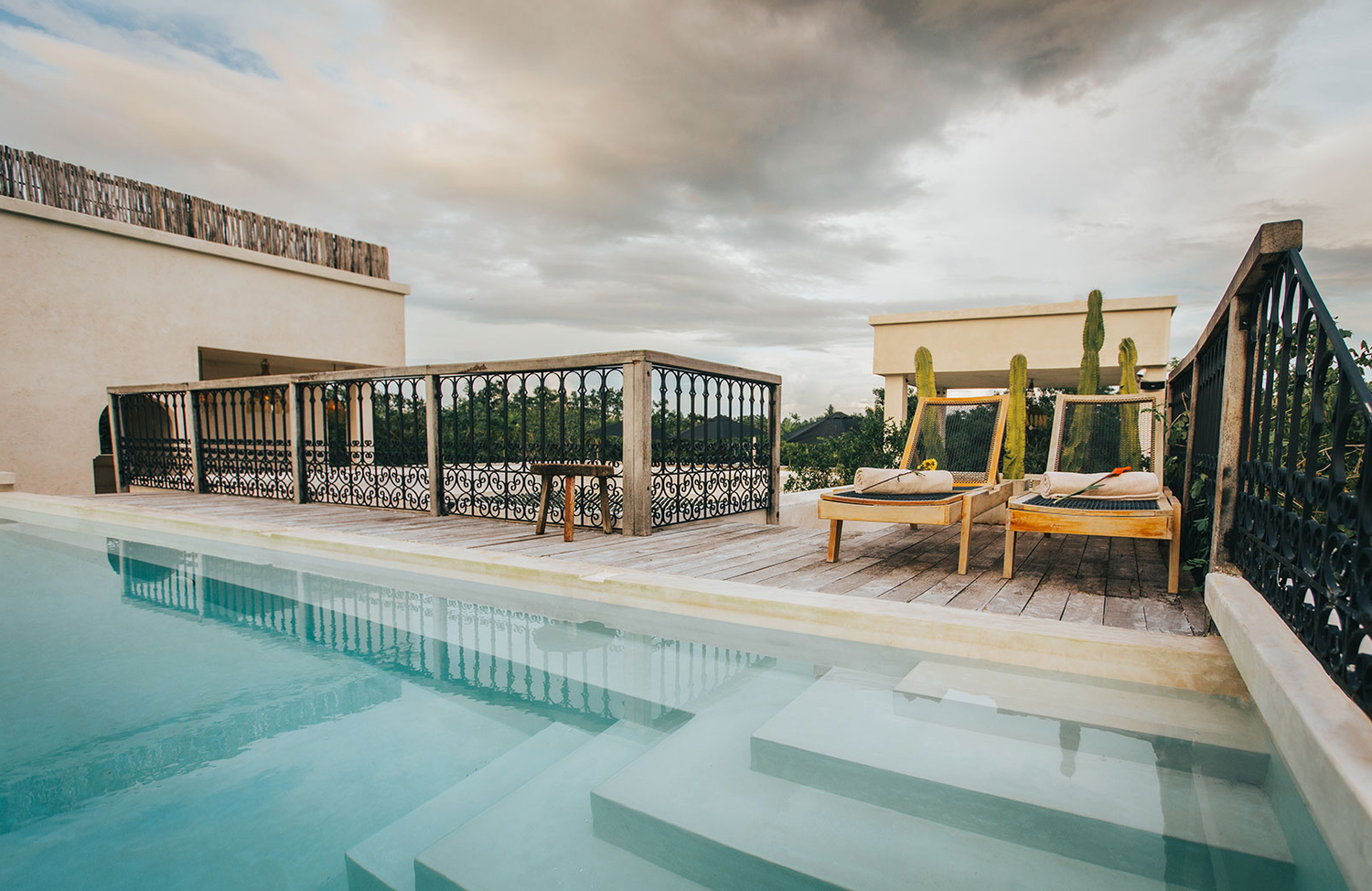 A modern rooftop pool deck features wooden loungers and cabanas under a cloudy sky.