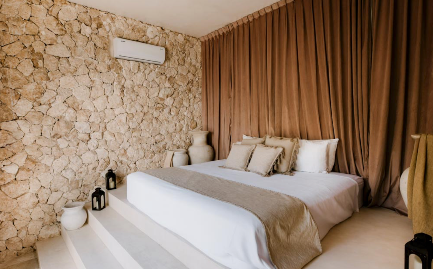 Modern bedroom interior with exposed stone walls, brown curtains, and white bedding in a luxury hotel setting.