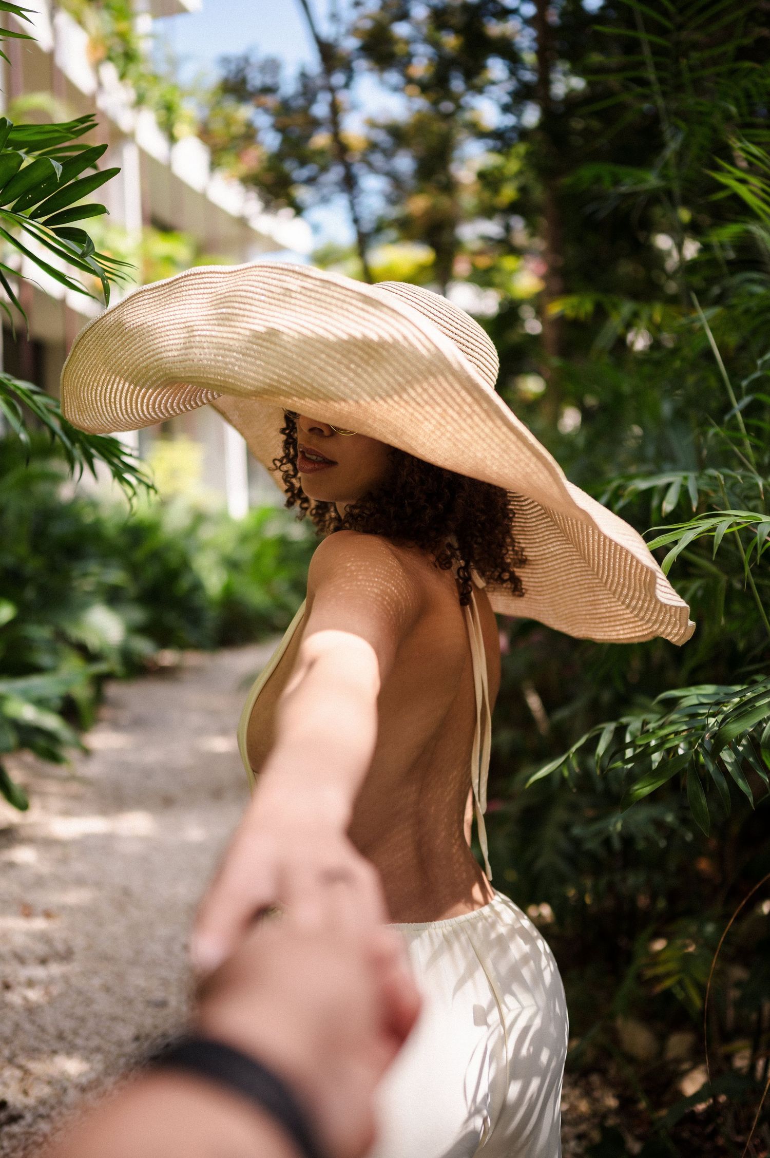 A figure wearing a wide-brimmed straw sun hat and white dress leads someone by the hand along a tropical garden path.