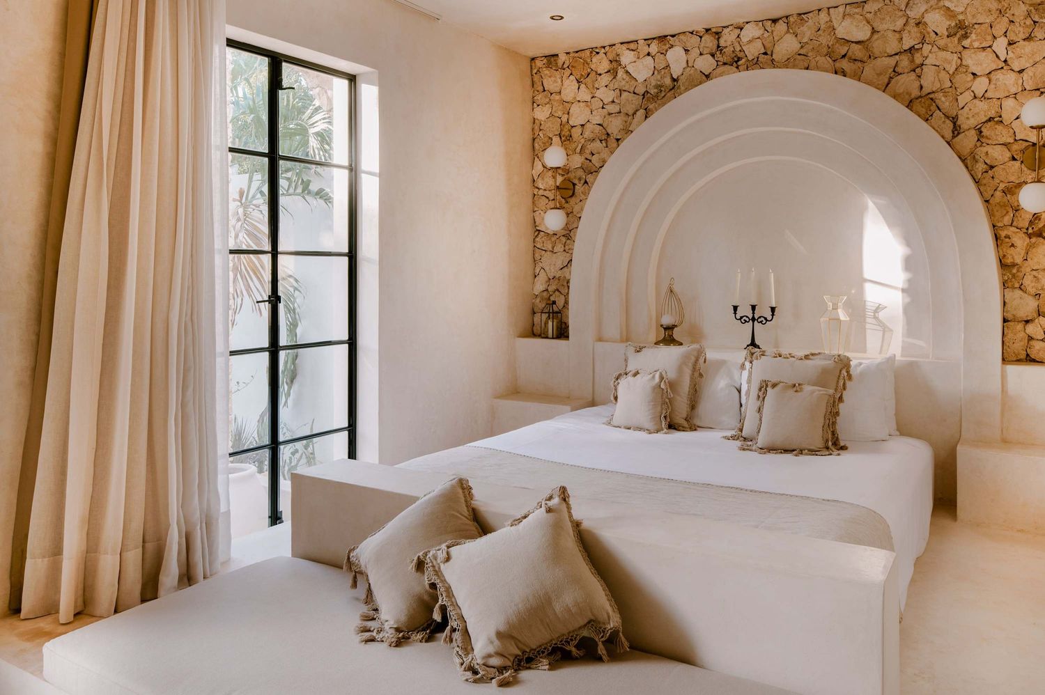 A luxurious Mediterranean-style bedroom with stone walls, arched doorways, and white bedding in natural light.
