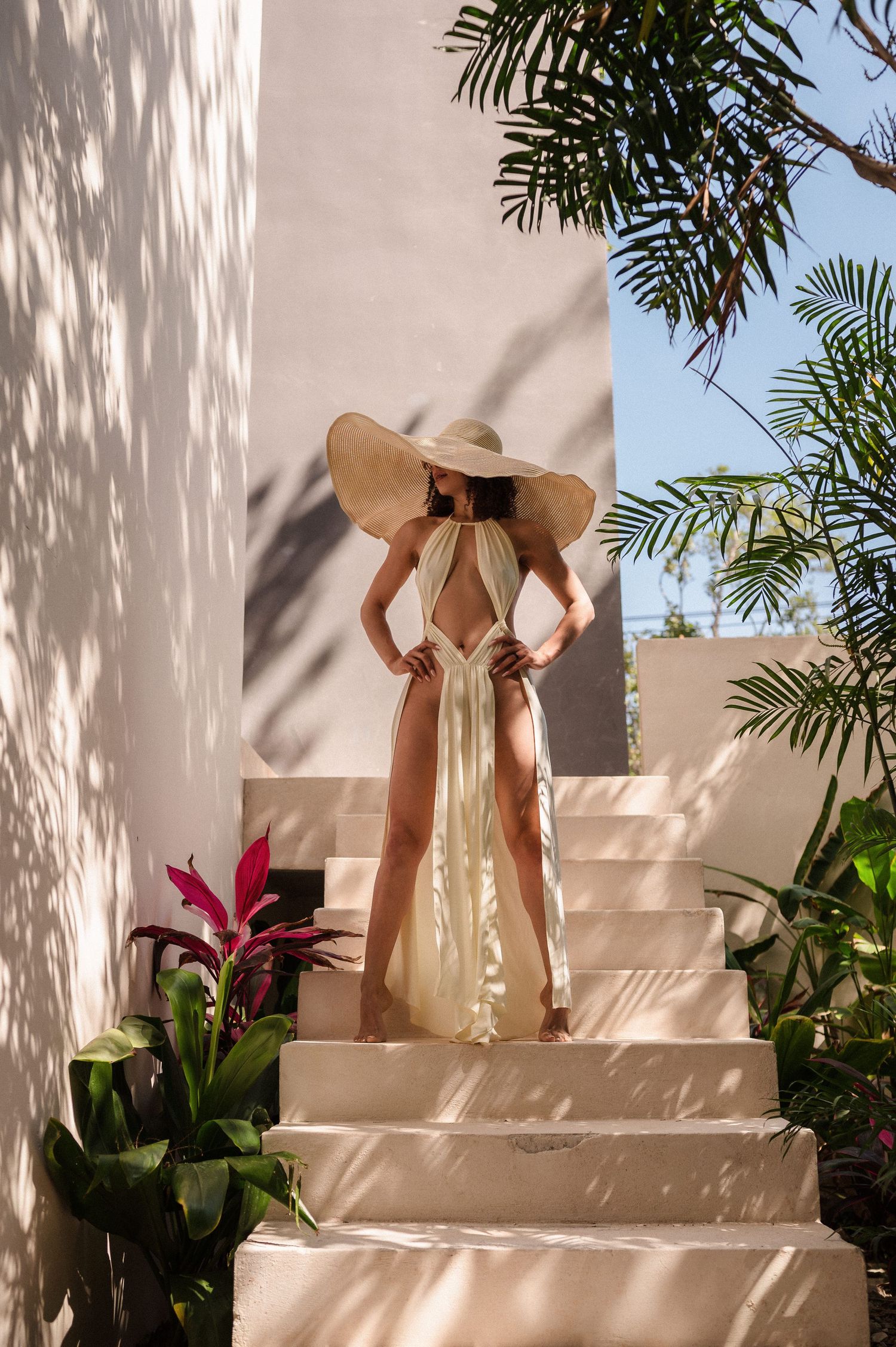 Figure in a wide-brimmed sun hat and flowing dress stands elegantly on white steps with tropical plants casting shadows.