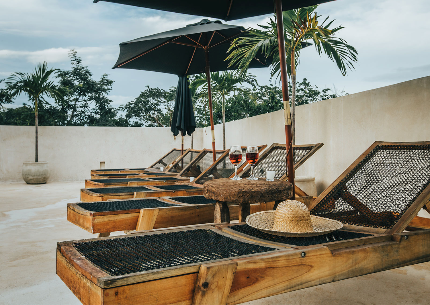 Wooden lounge chairs with gray cushions arranged on a terrace overlooking palm trees and a cloudy sky.