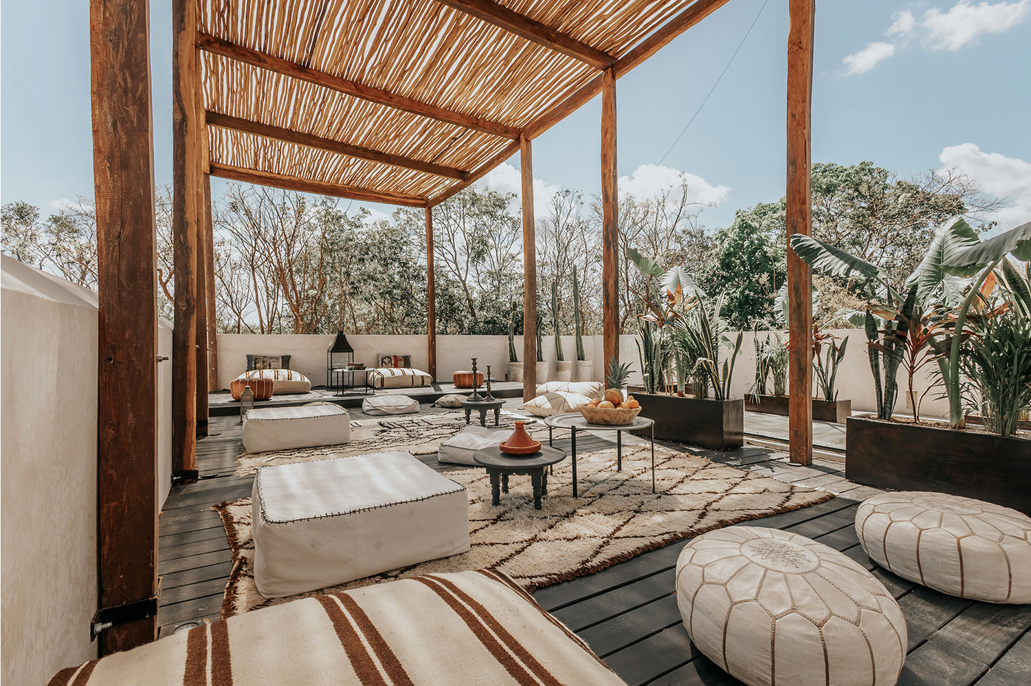 Modern outdoor lounge area with wooden pergola, plush cushions and poufs on a wooden deck under natural sunlight.