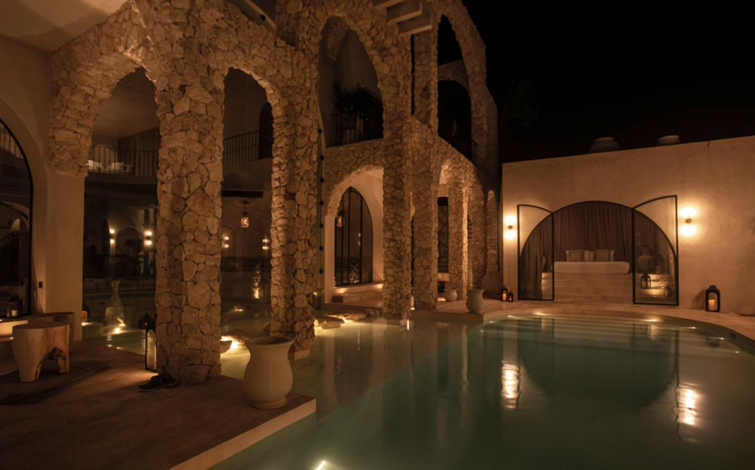 Illuminated stone archways reflect on still water in a luxurious indoor pool at night.