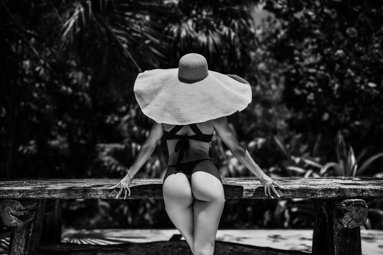 Black and white photo of a person in a large sun hat sitting on a wooden railing with their back to the camera.