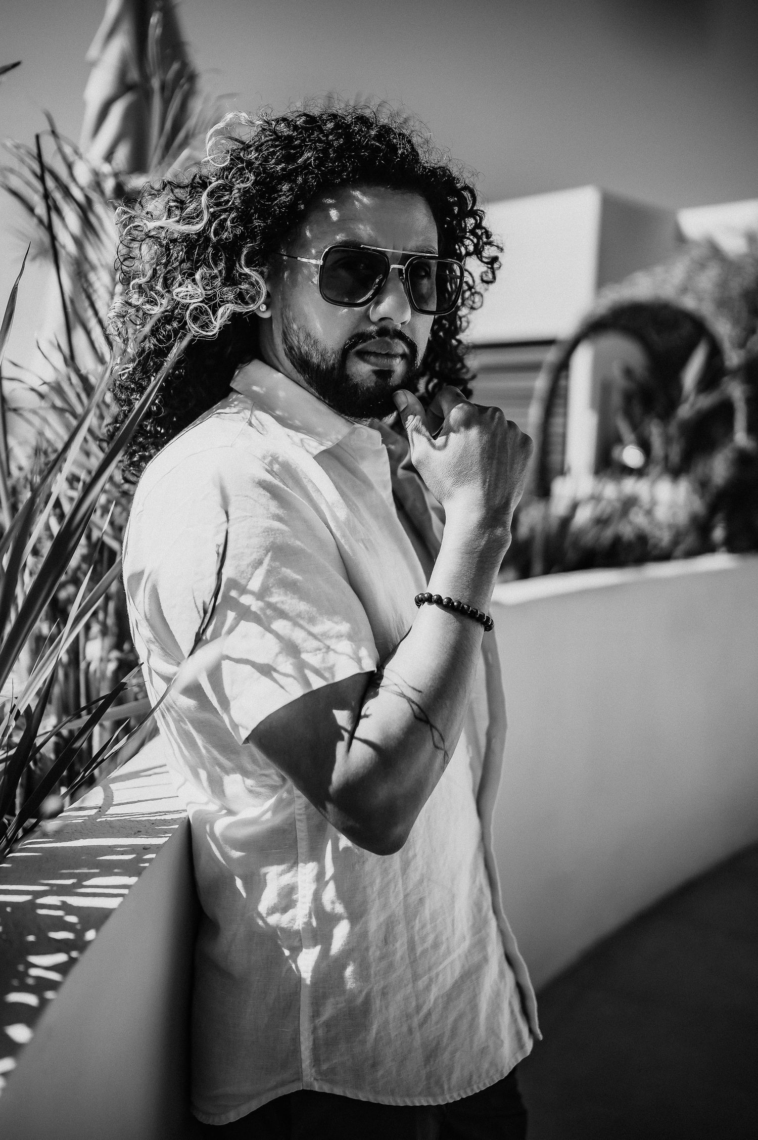 Black and white portrait of person in sunglasses and white shirt with curly hair standing by wall in natural light.