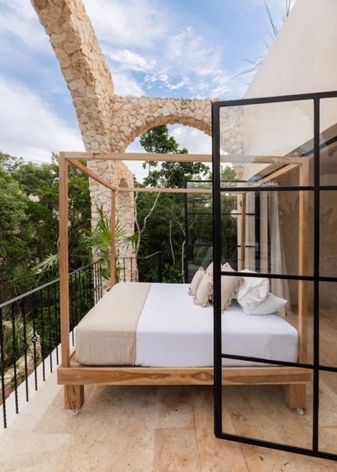 Luxurious outdoor bed with white linens on wooden frame beneath stone archway and glass doors overlooking greenery.