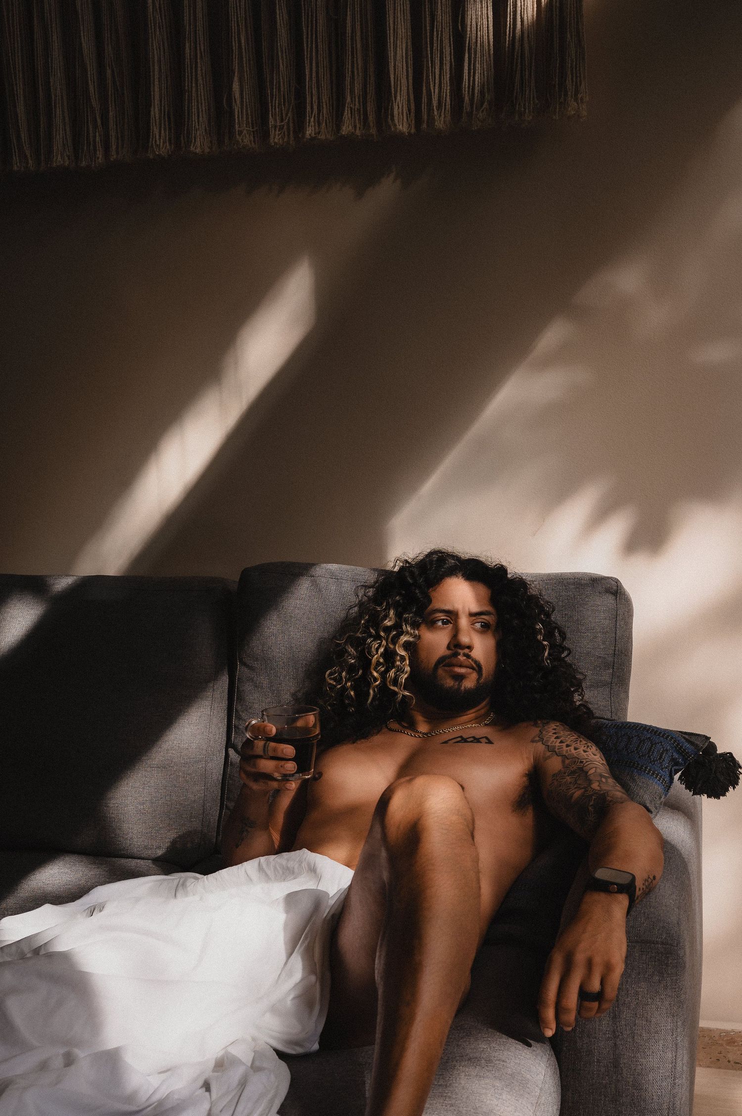 Artistically lit indoor portrait showing shirtless figure with curly hair lounging on grey couch with white sheets.