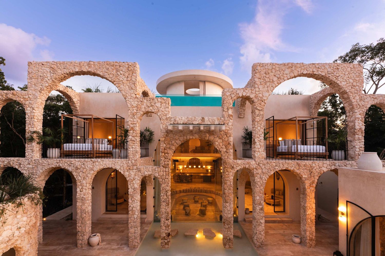 Ancient stone arches and columns illuminate a rustic Mediterranean courtyard at dusk with warm glowing lights.