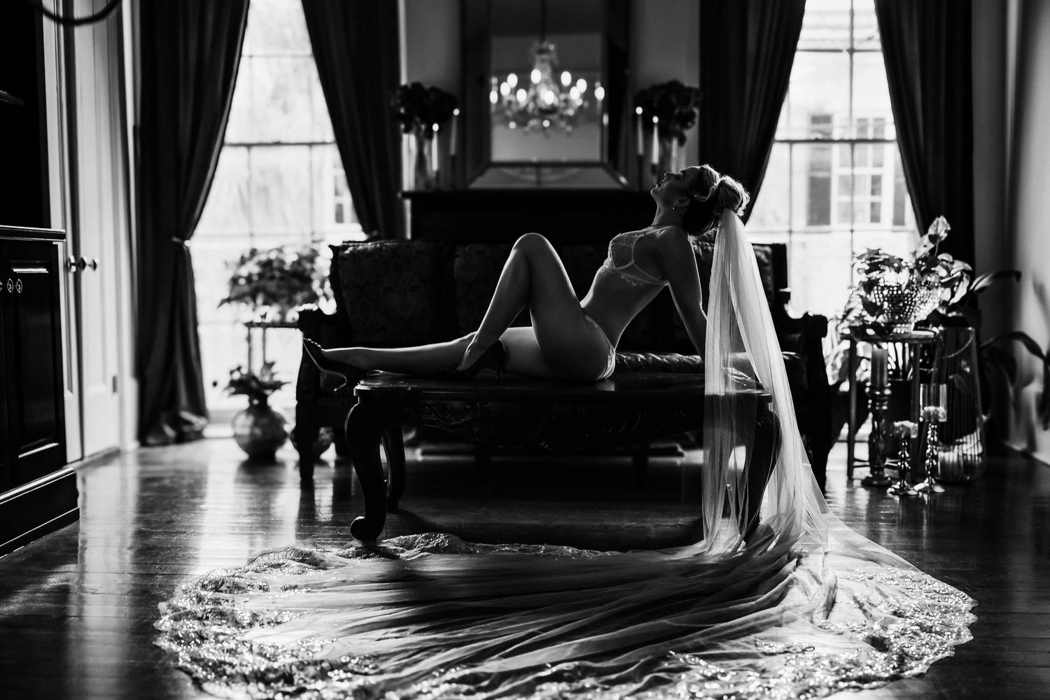 Black and white boudoir photo of a bride in lingerie and veil reclining elegantly on a vintage sofa in a grand room.