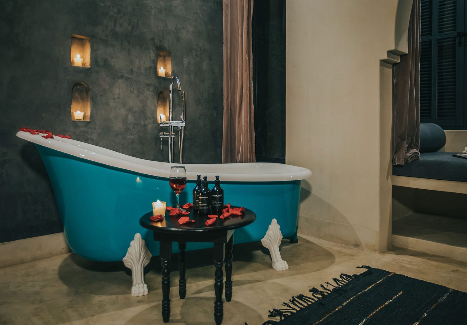 A vintage turquoise clawfoot bathtub with candles and a wooden side table in a moody, spa-like bathroom setting.