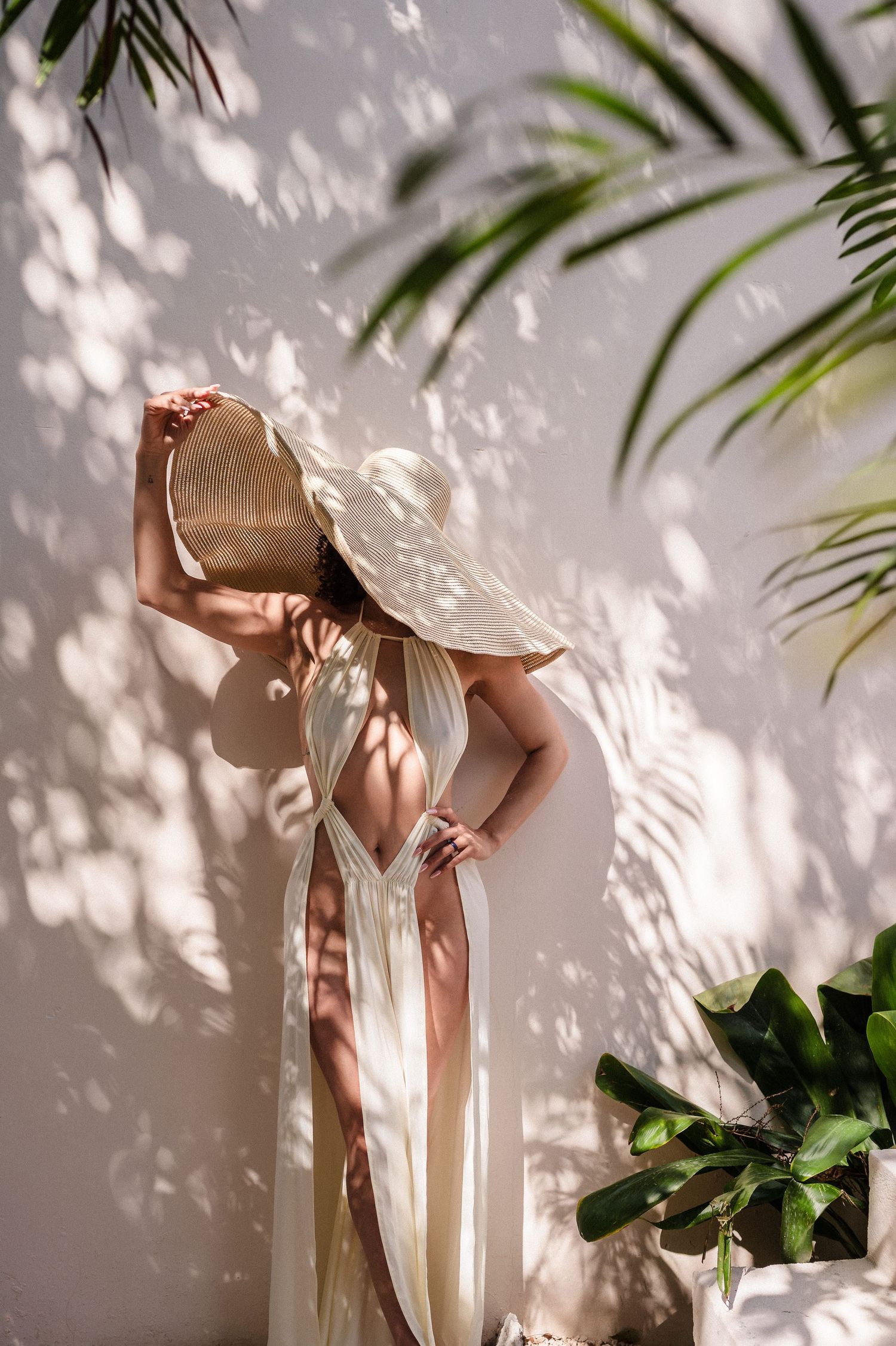 Shadows from palm leaves create dappled patterns on a wall while a figure in white stands gracefully in natural light.