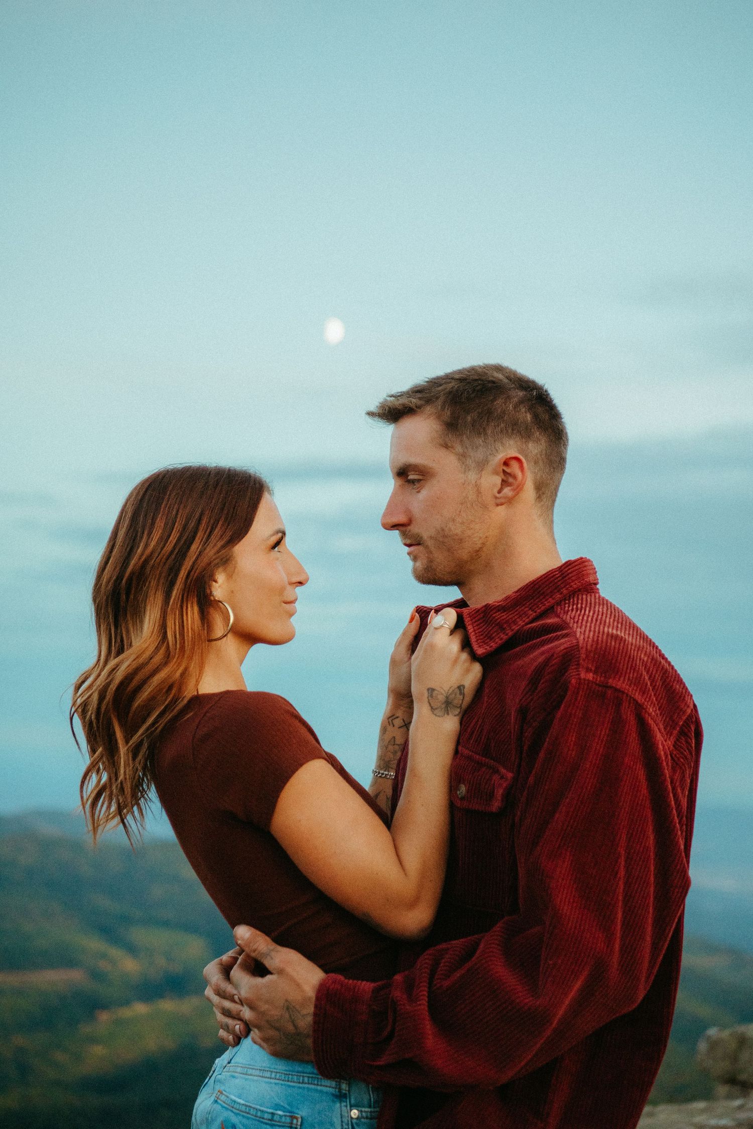 A romantic couple embraces on a mountaintop at dusk with a moon visible in the pale blue sky.