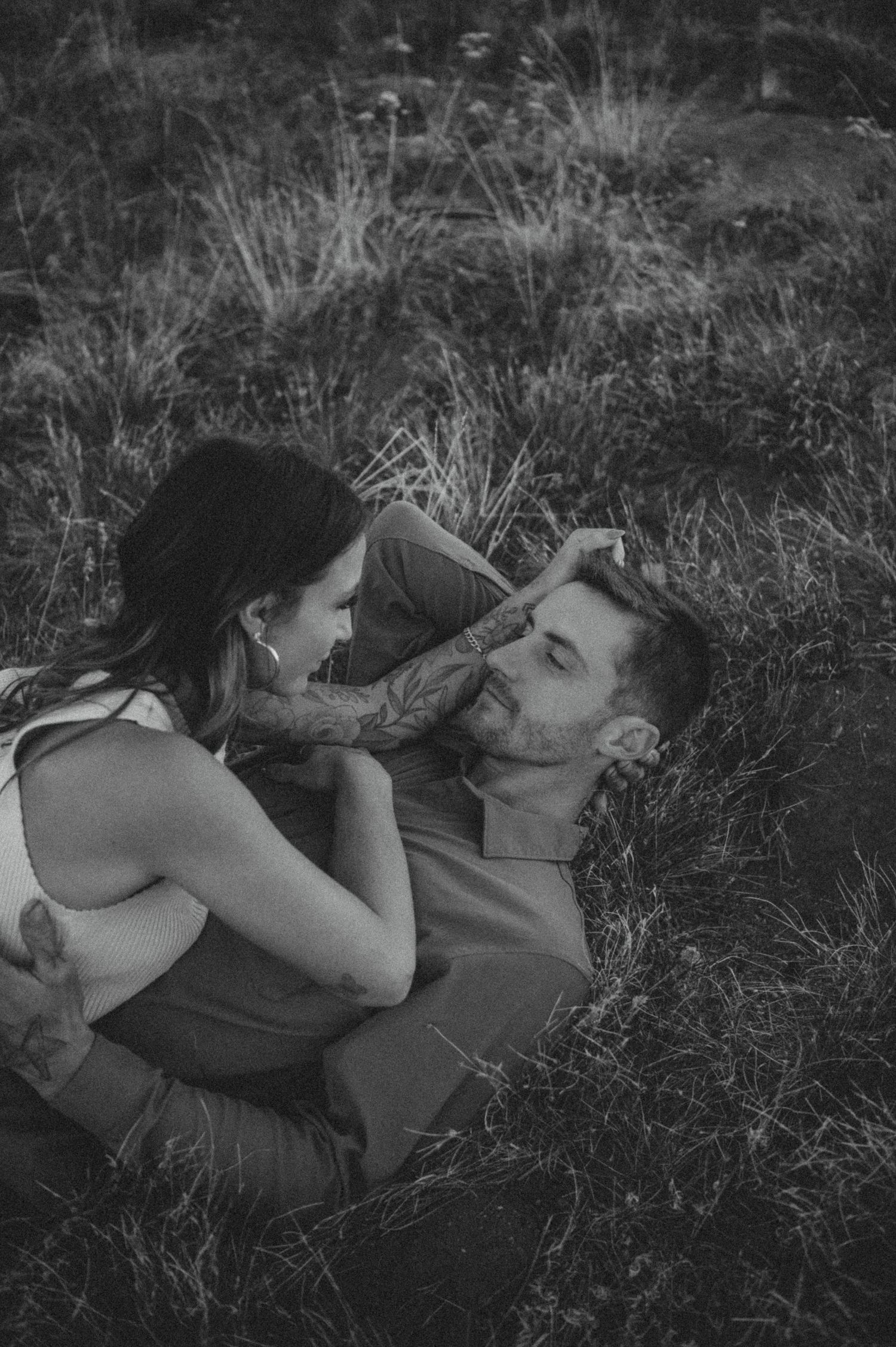 A tender black and white photo shows a couple laying in tall grass, sharing an intimate moment together.