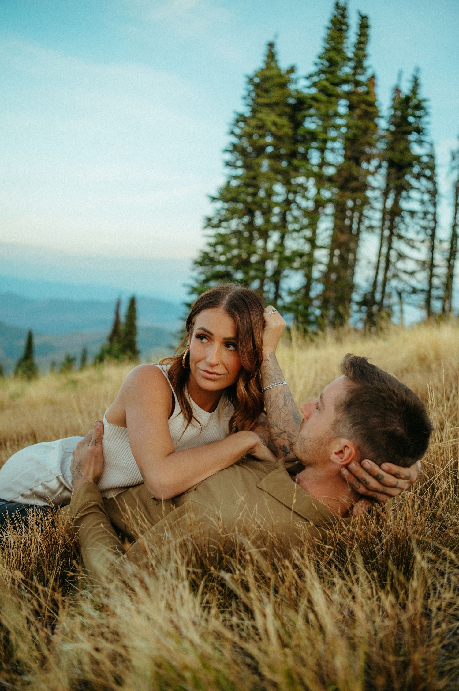 A couple laying in golden grass with tall evergreen trees and mountain landscape during sunset.