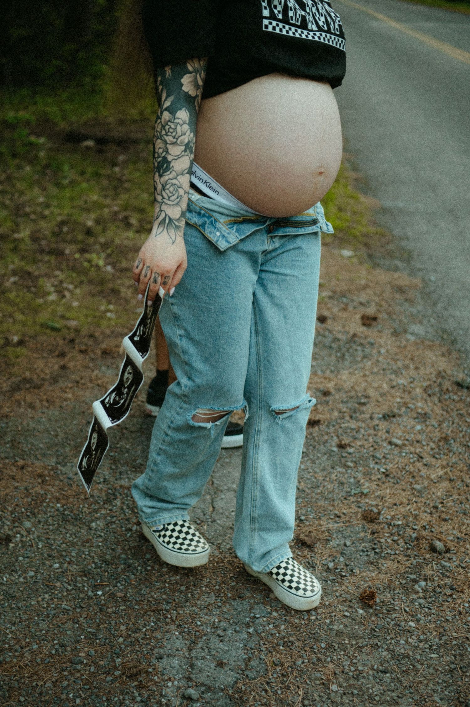 A pregnant person in ripped jeans and checkered Vans holds a camera while standing on a gravel road.