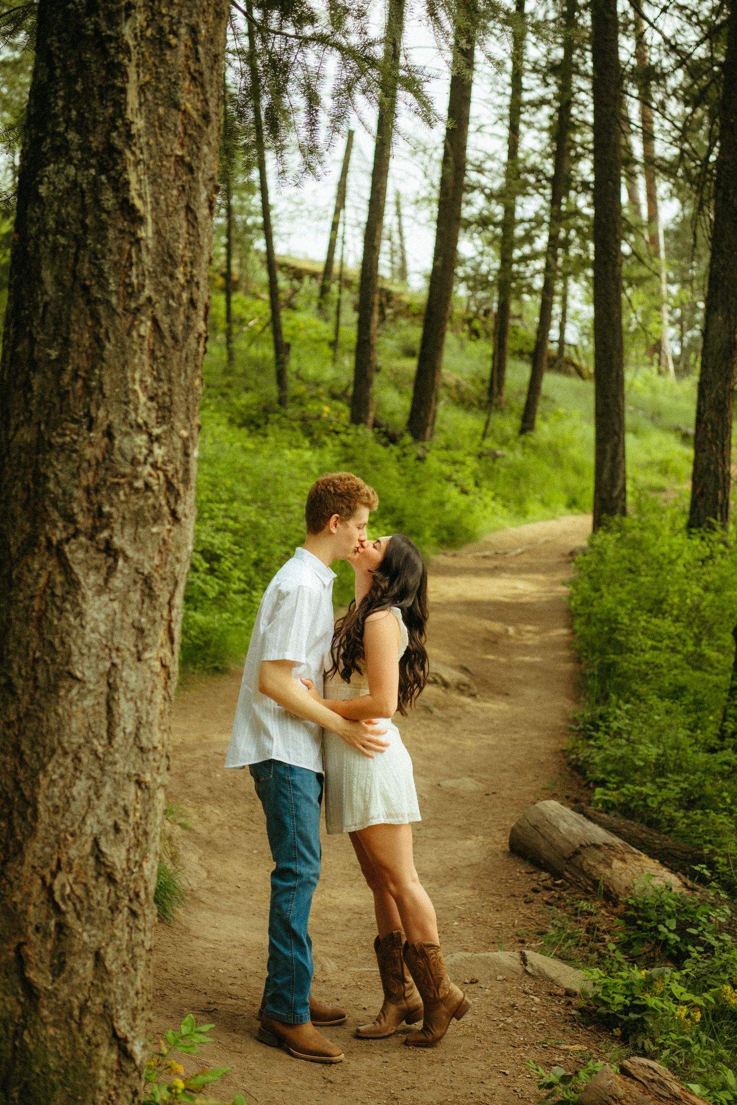 A romantic couple embraces on a winding forest trail surrounded by tall evergreen trees and lush greenery.