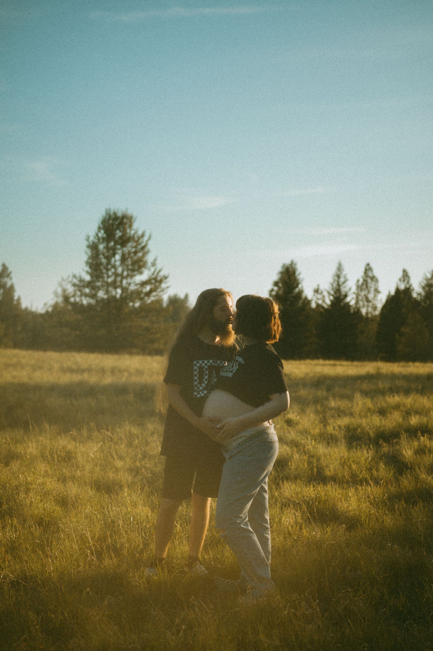 A couple embraces in a sunlit meadow at golden hour with evergreen trees silhouetted in the background.