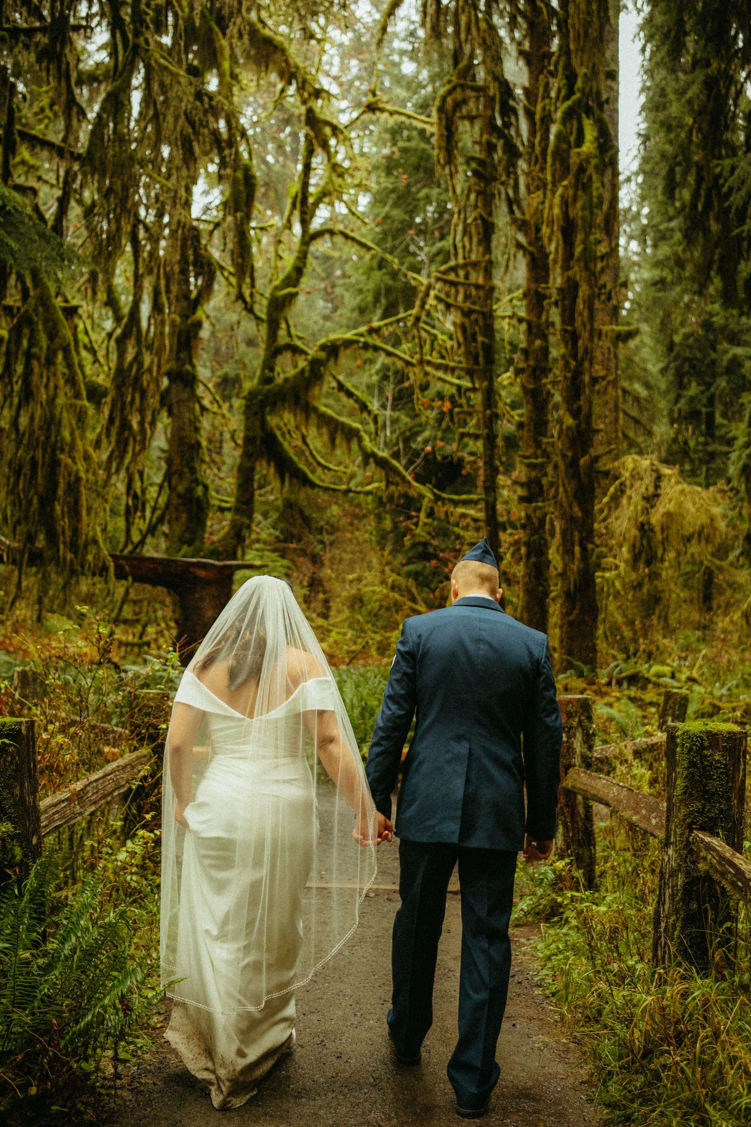A couple in wedding attire walks down a mossy forest path surrounded by towering evergreen trees draped in moss.