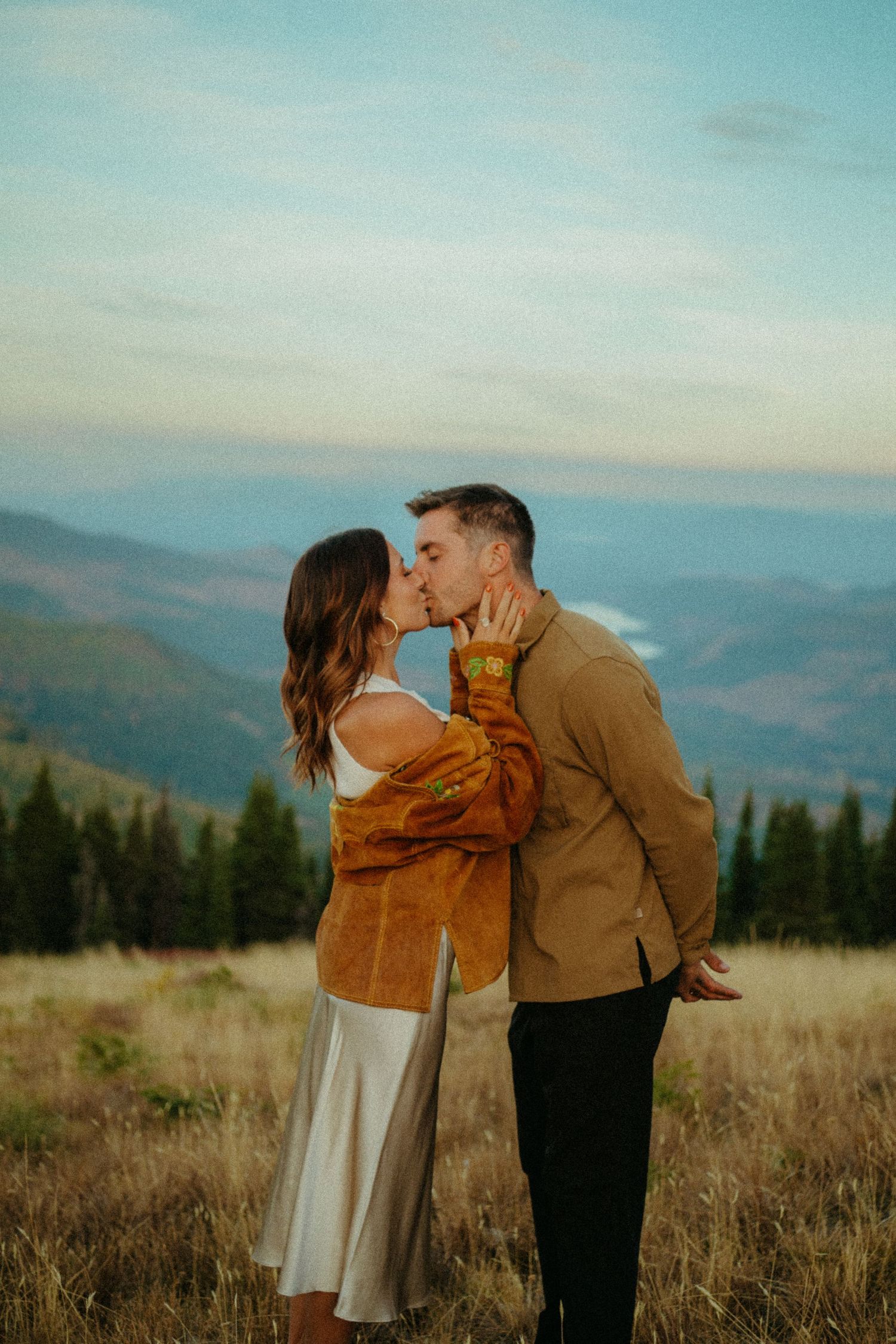 Romantic couple sharing an intimate moment at sunset in a scenic mountain landscape overlooking distant hills and forests.