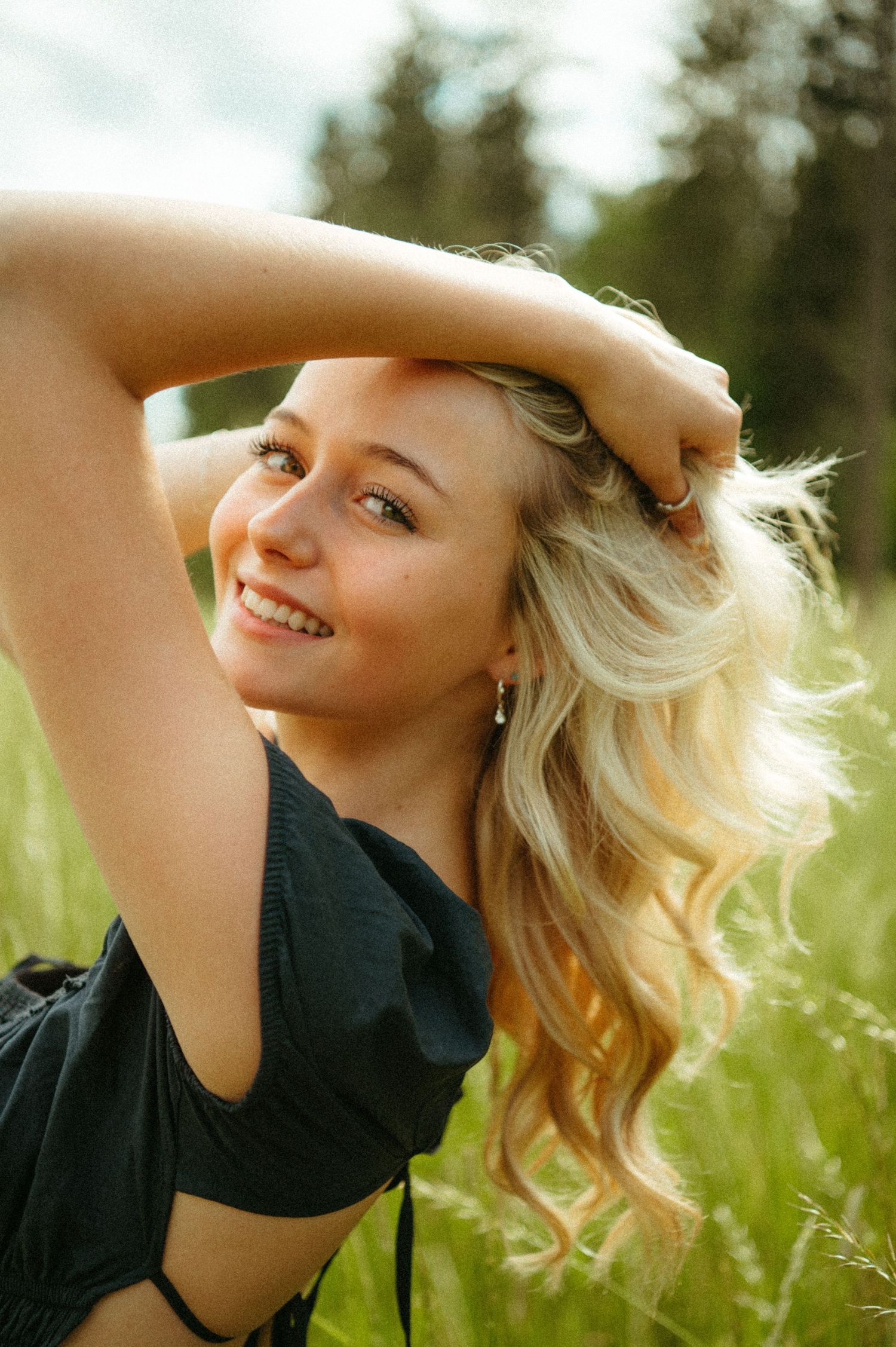 Person with blonde hair poses in a black shirt against a sunlit natural background with tall grass and greenery.