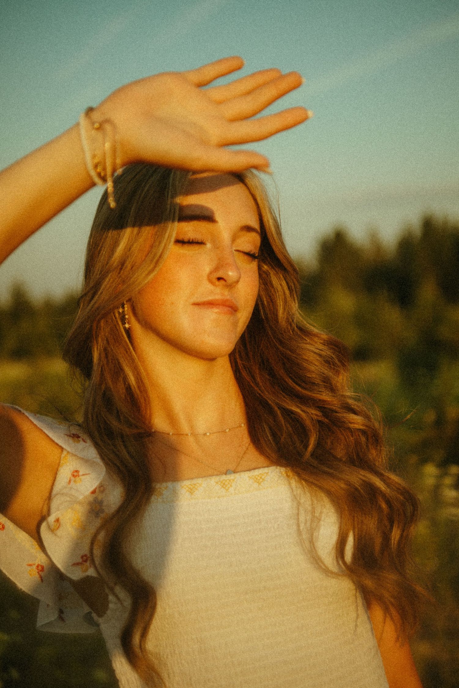 A dreamy portrait of someone with long wavy hair wearing white in golden sunlight with an upraised hand.