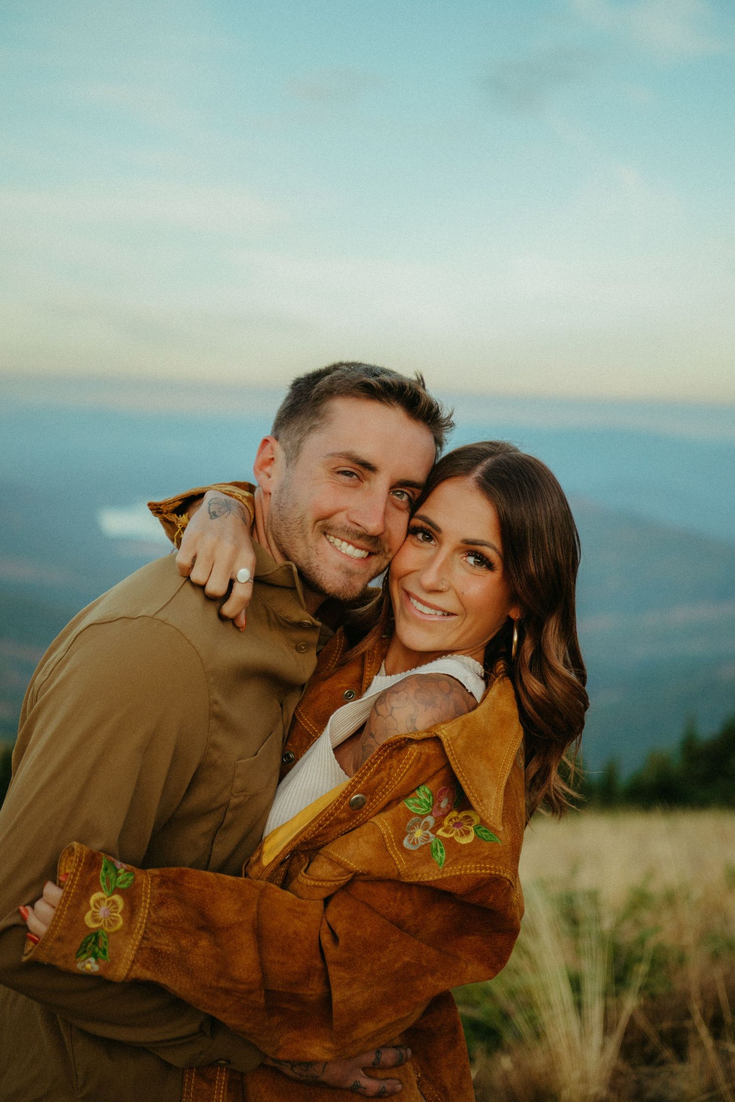 A couple embraces outdoors wearing fall jackets against a scenic mountain backdrop at sunset.
