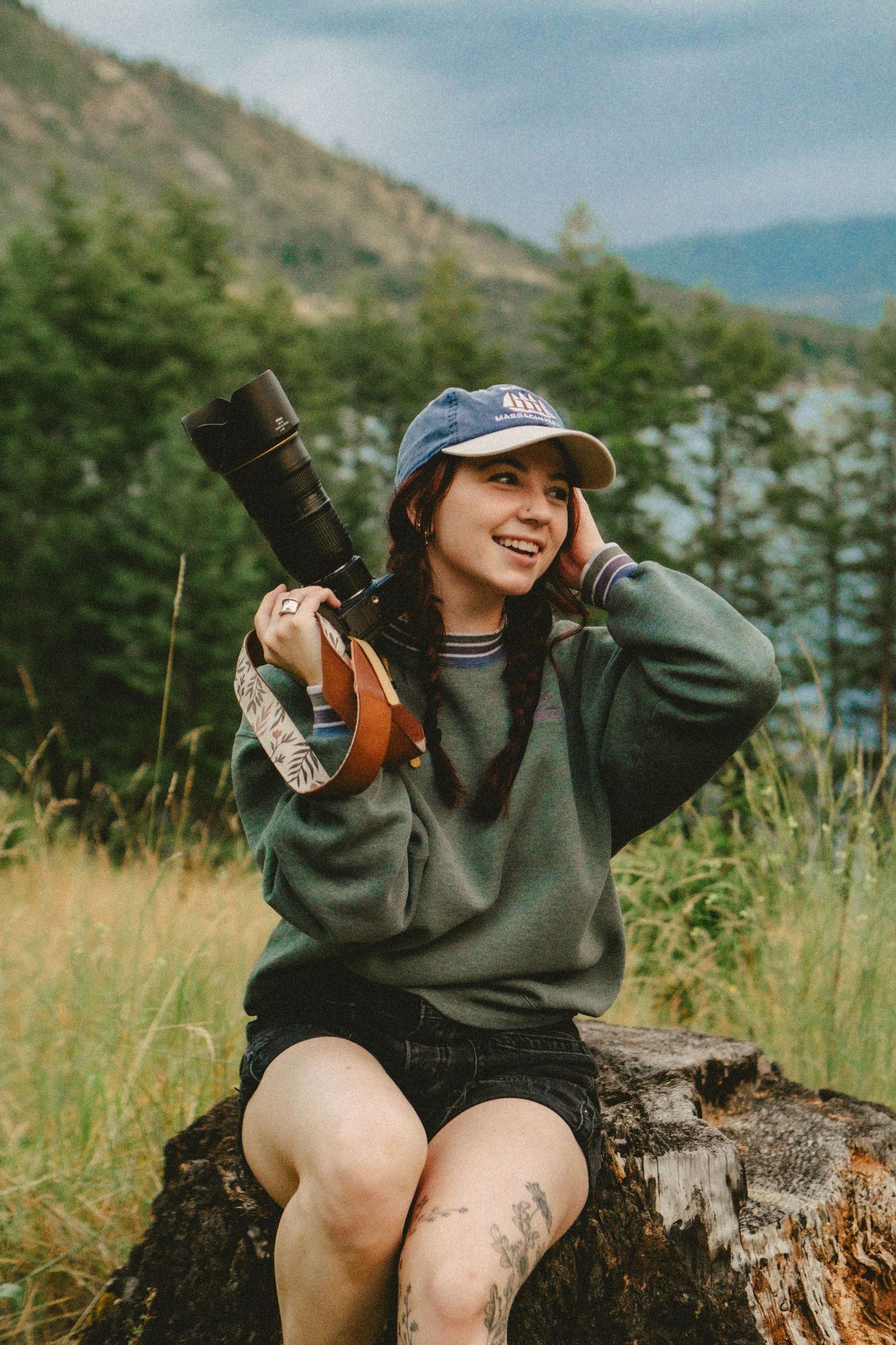 Nature photographer in casual attire sits on tree stump with camera and telephoto lens against mountain landscape.