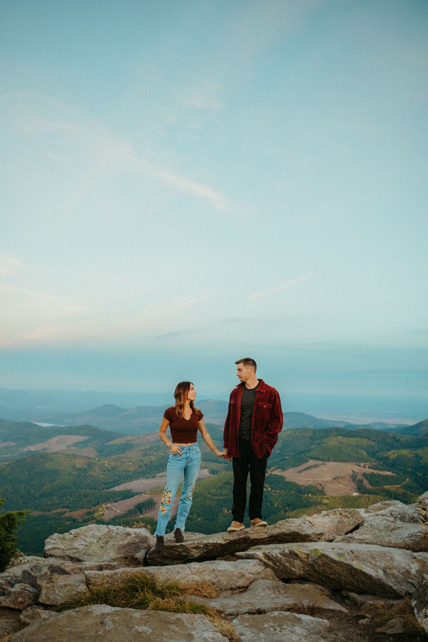 A couple stands together on a rocky mountain summit overlooking a scenic valley landscape at sunset.