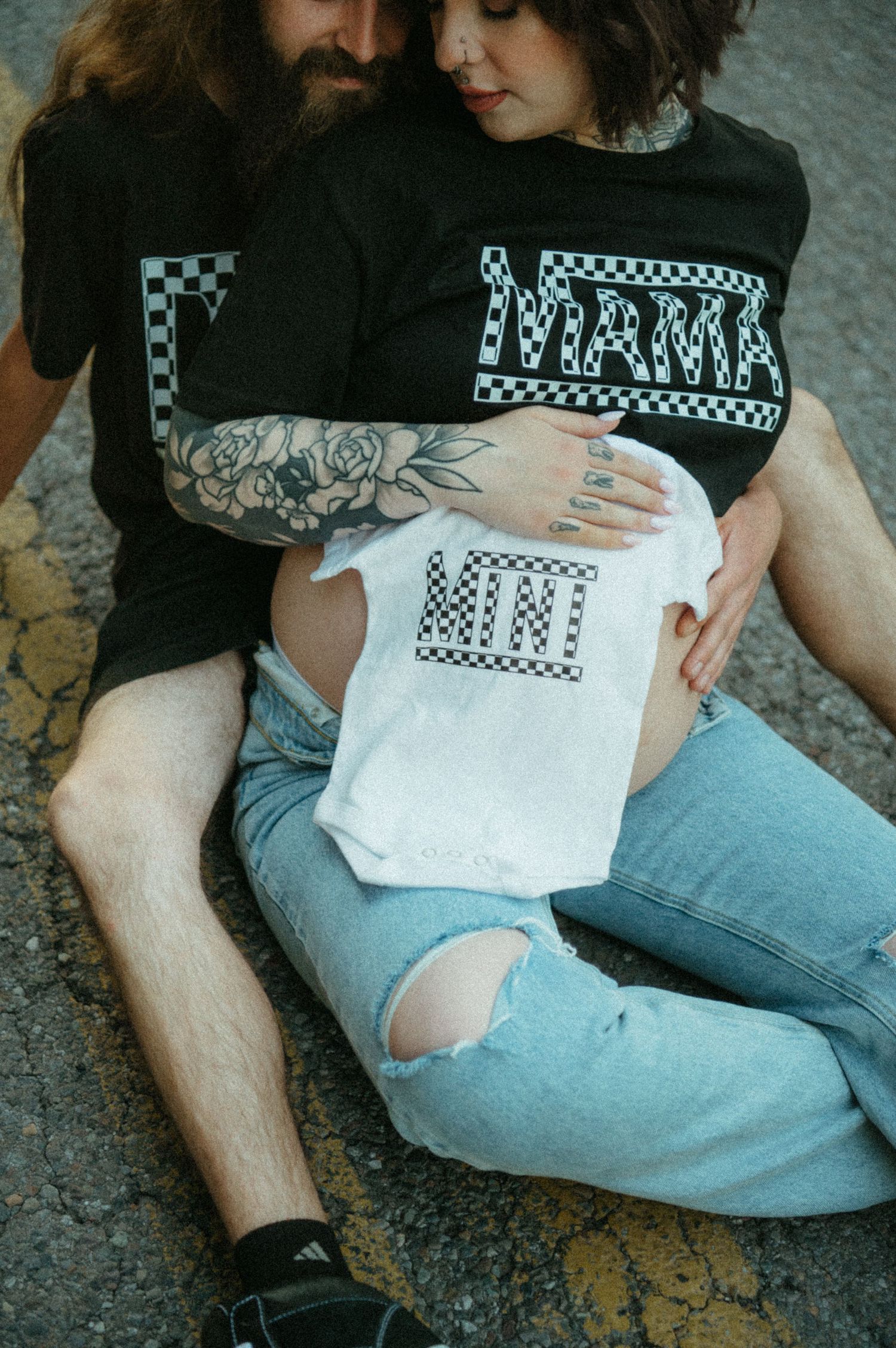 Couple wearing matching black and white checker-print shirts showing off a baby onesie with similar design on pavement.