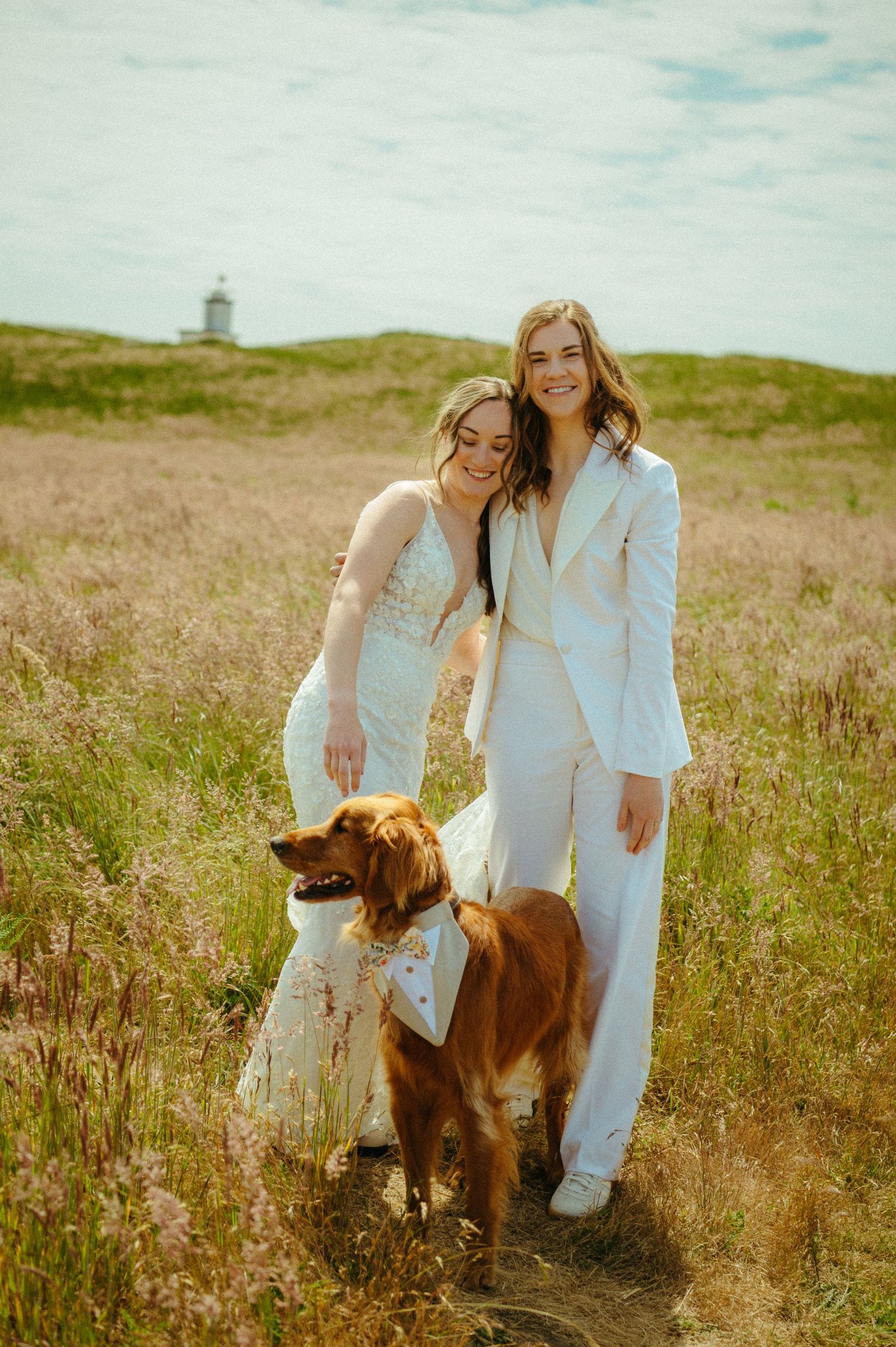 Two people in white outfits pose with their brown dog in a grassy field with a lighthouse visible in the background.