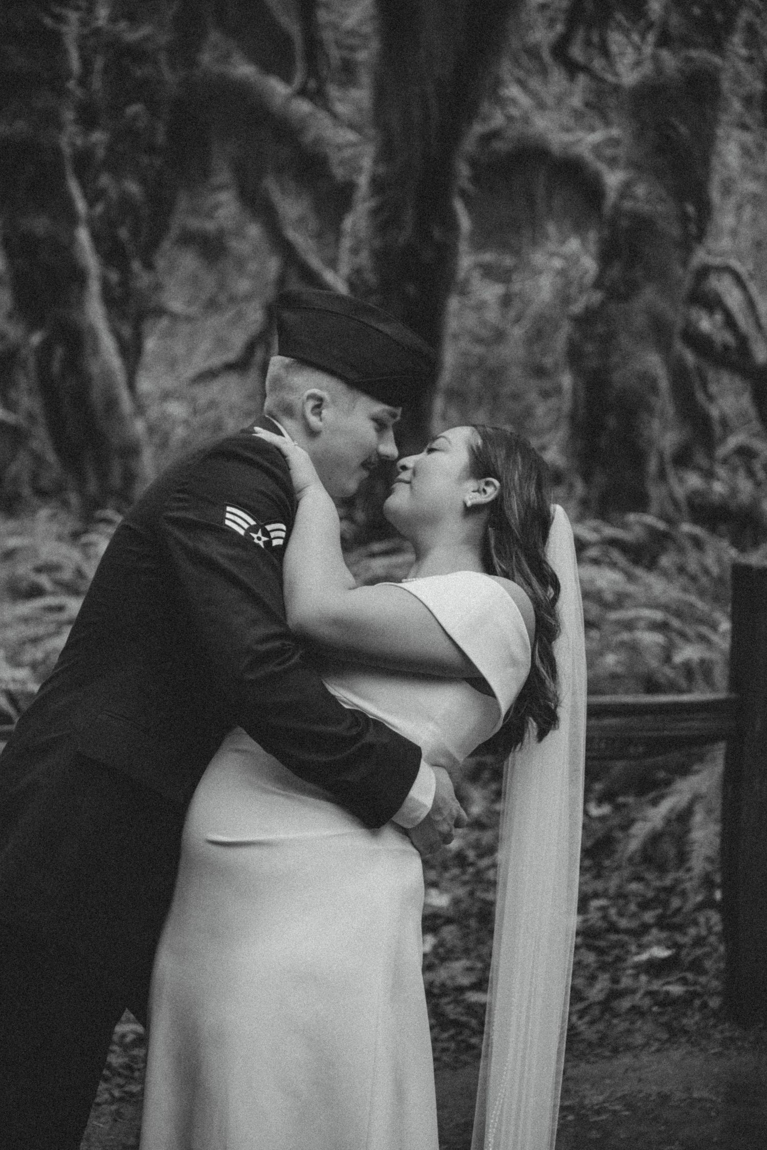 Black and white wedding photo capturing a romantic embrace between a bride in white dress and military groom in uniform outdoors.